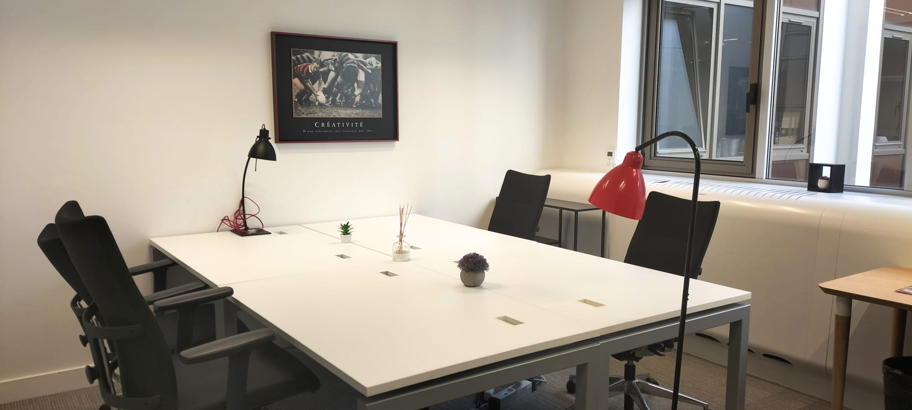 The image shows a modern office meeting room with a white table, four black chairs, a red desk lamp, and a small plant on the table, along with a framed artwork on the wall.