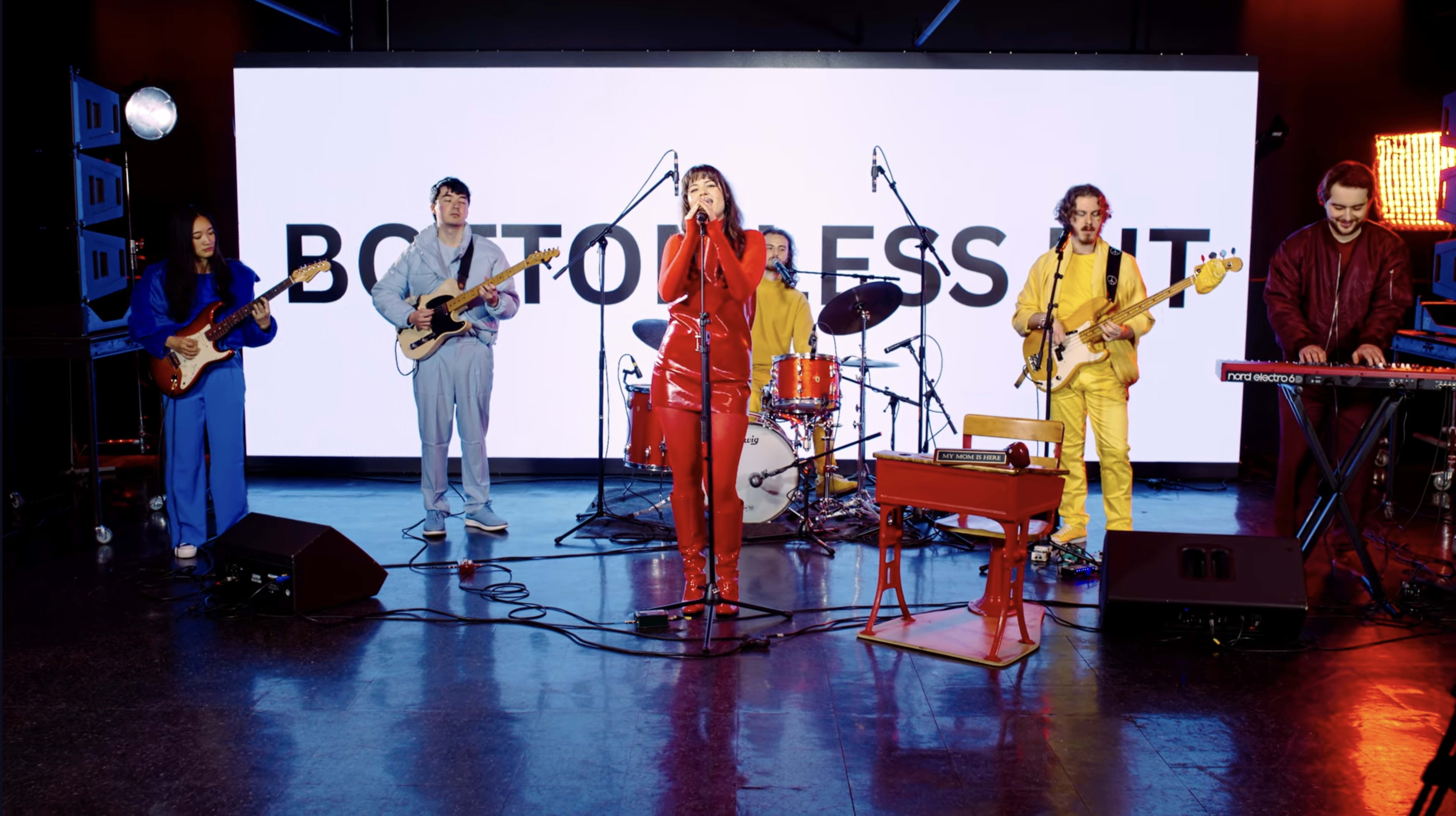 A band performs on stage with various instruments and colorful outfits in front of a large screen displaying the text "BOTTOM DRESS IT."