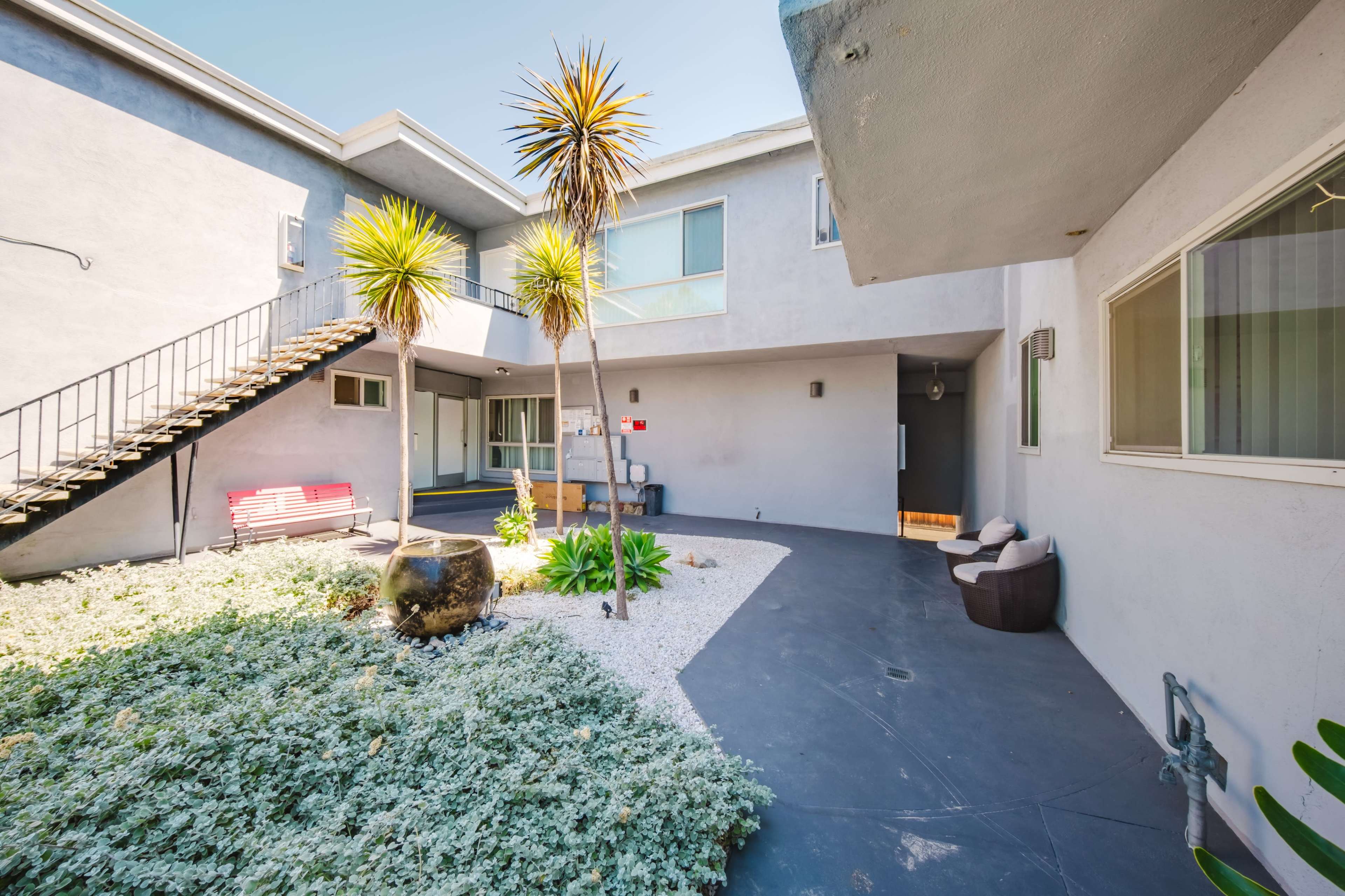 The image shows a modern courtyard area of an apartment complex featuring palm plants, gravel landscaping, and a staircase leading to the upper level.