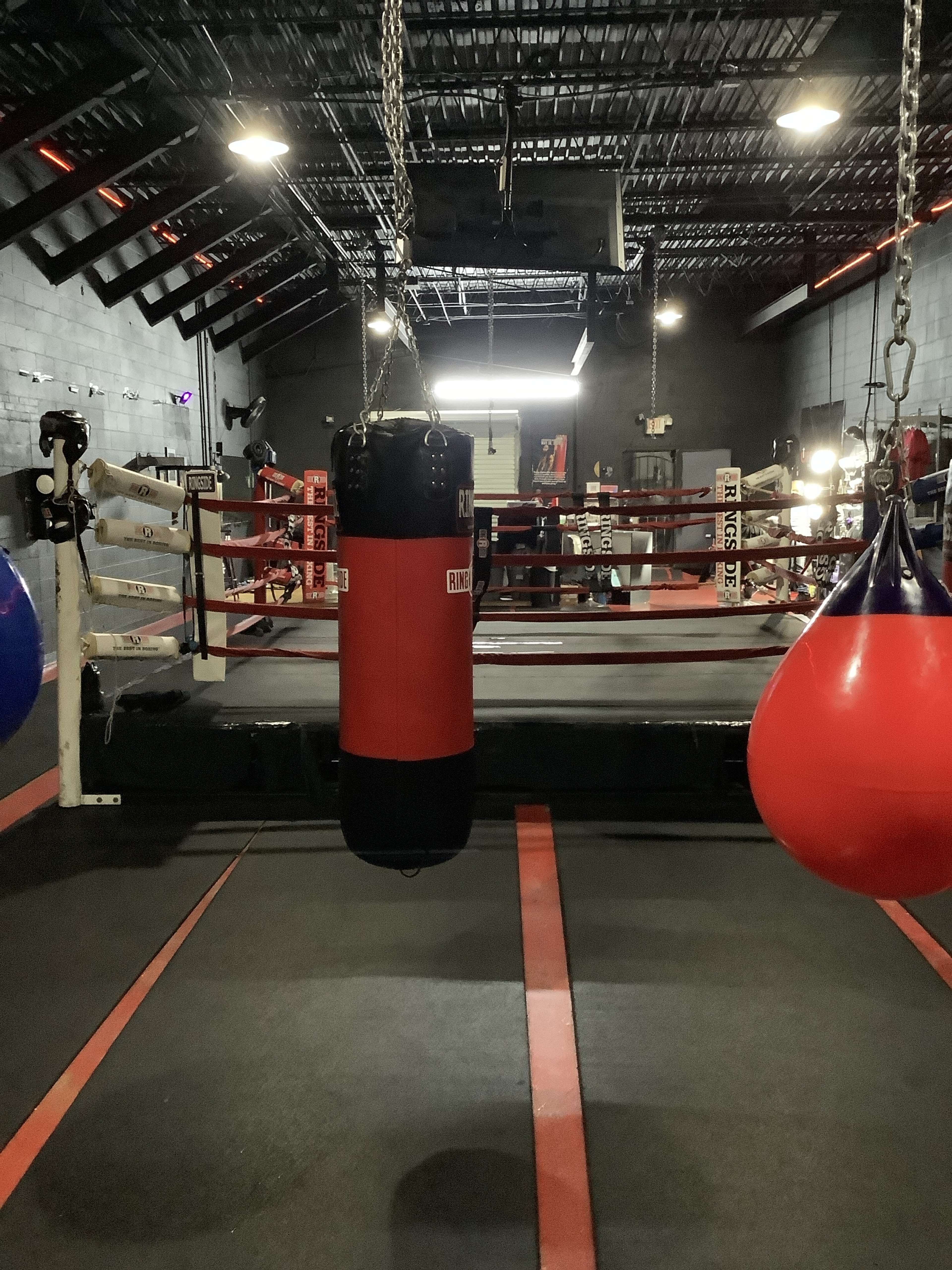 A boxing gym with a red punching bag suspended from the ceiling, surrounded by a boxing ring and exercise equipment.