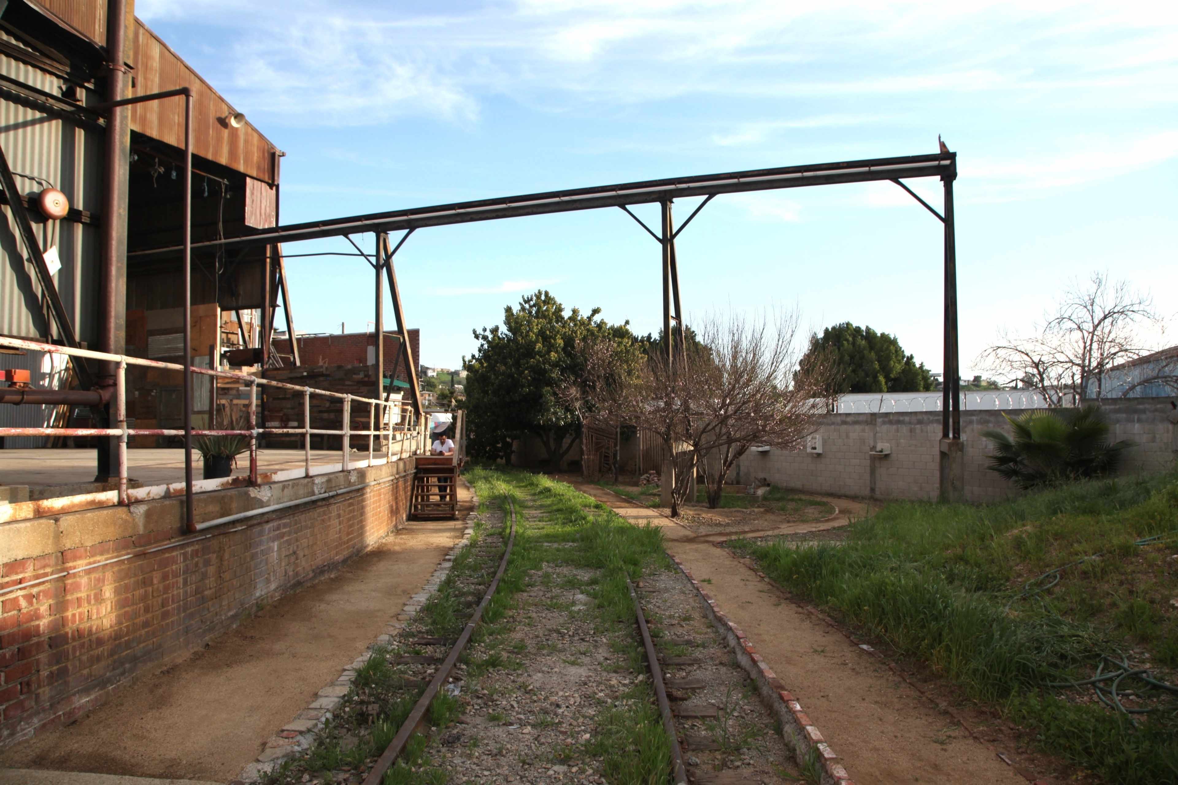 The image shows an abandoned industrial area with rusty train tracks running alongside a building and overgrown grass on either side.