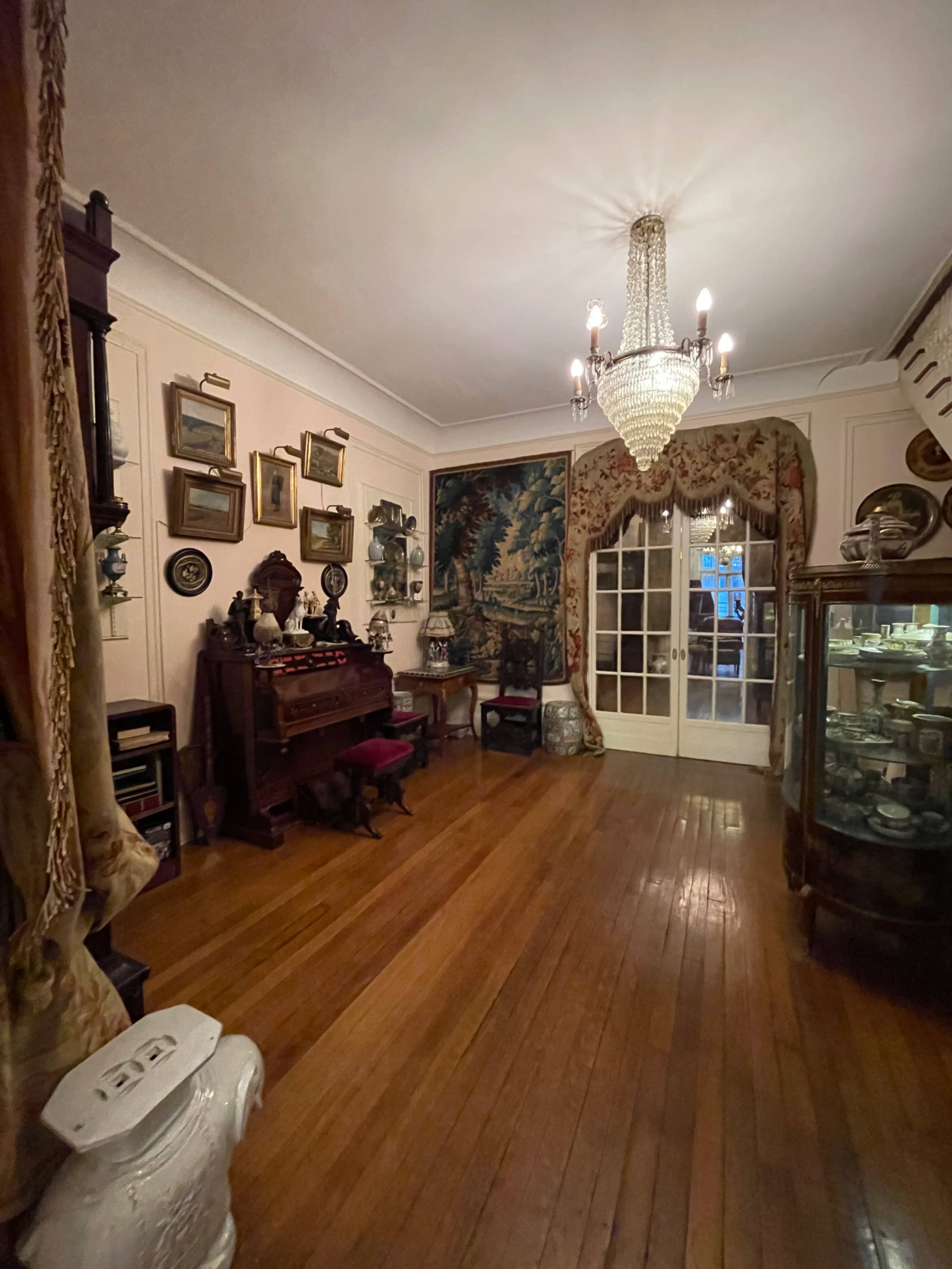 The image shows a vintage room with wooden floors, ornate furniture, a crystal chandelier, and a large tapestry on the wall, alongside various decorative items displayed in a glass cabinet.