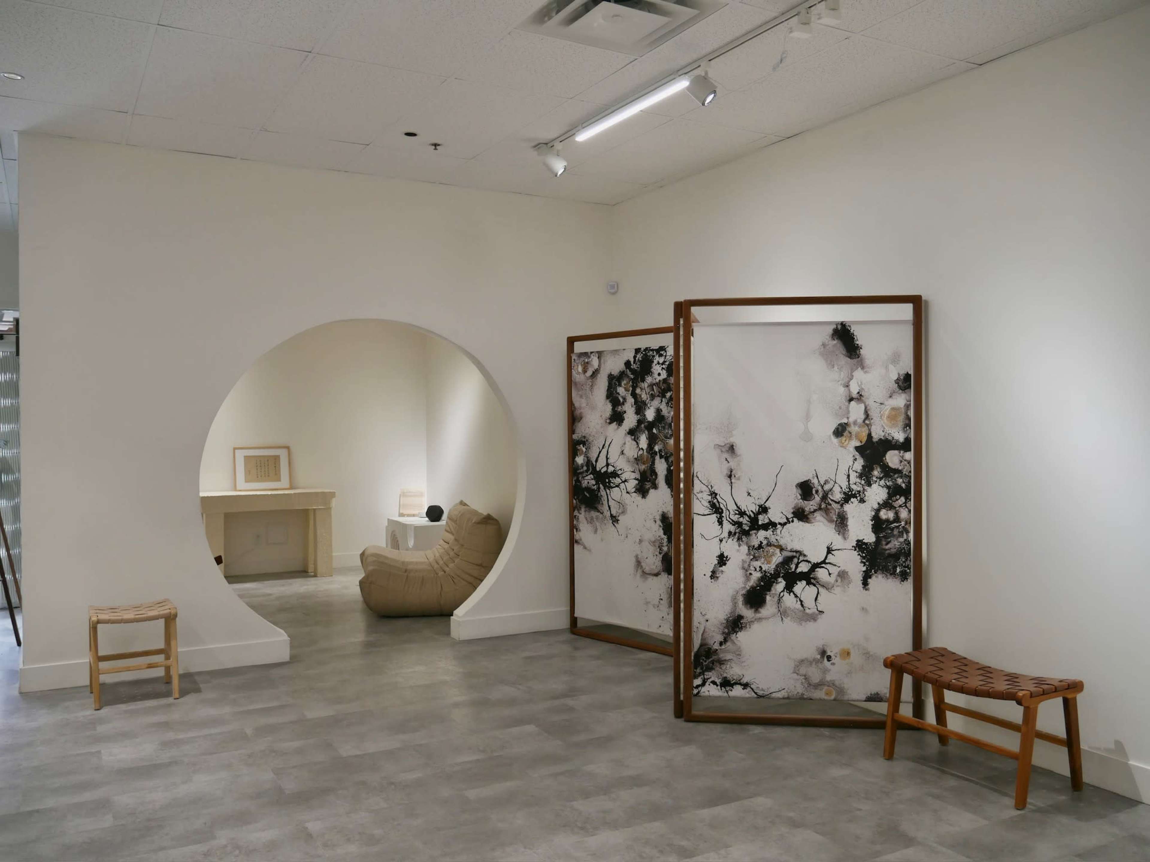 A minimalist room featuring a curved archway leading to a beige chair, a wooden folding screen with a black and white design, and two wooden benches against light-colored walls and flooring.