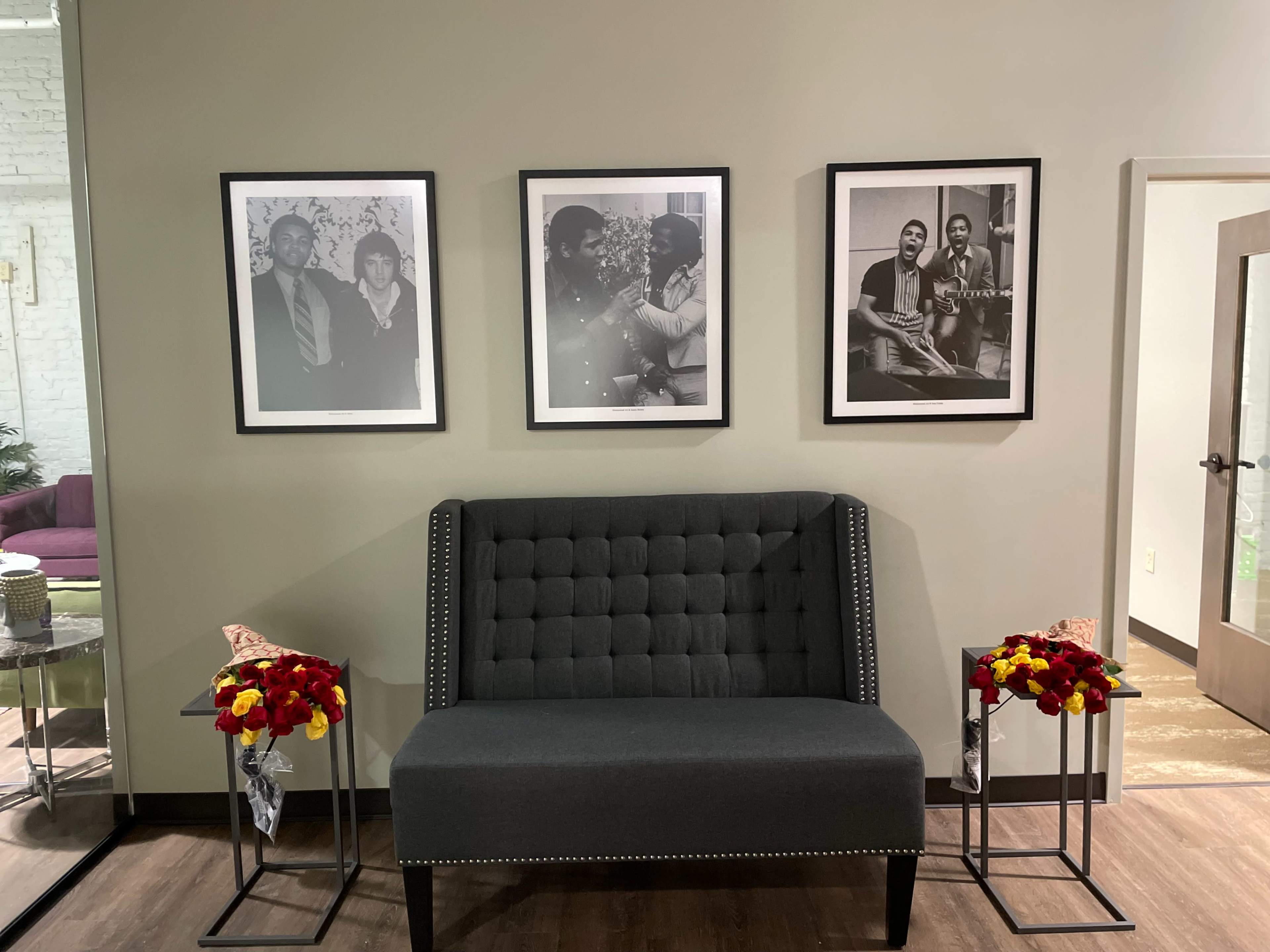 A gray upholstered bench is centered against a wall adorned with four black-and-white framed photographs, with two floral arrangements on small stands on either side.