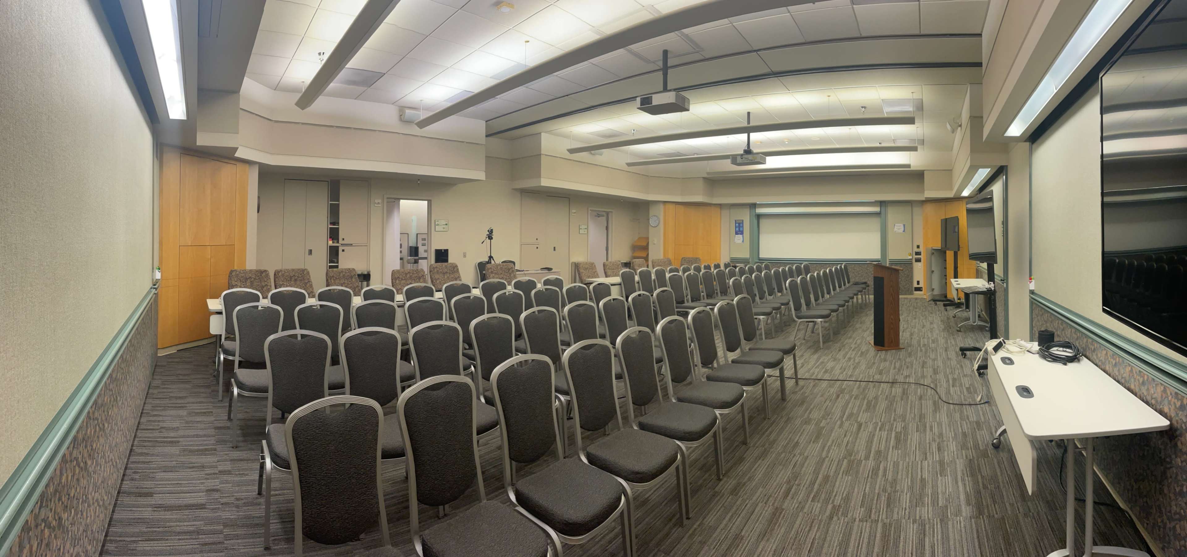 A panoramic view of a conference room with rows of empty chairs facing a podium and a screen.