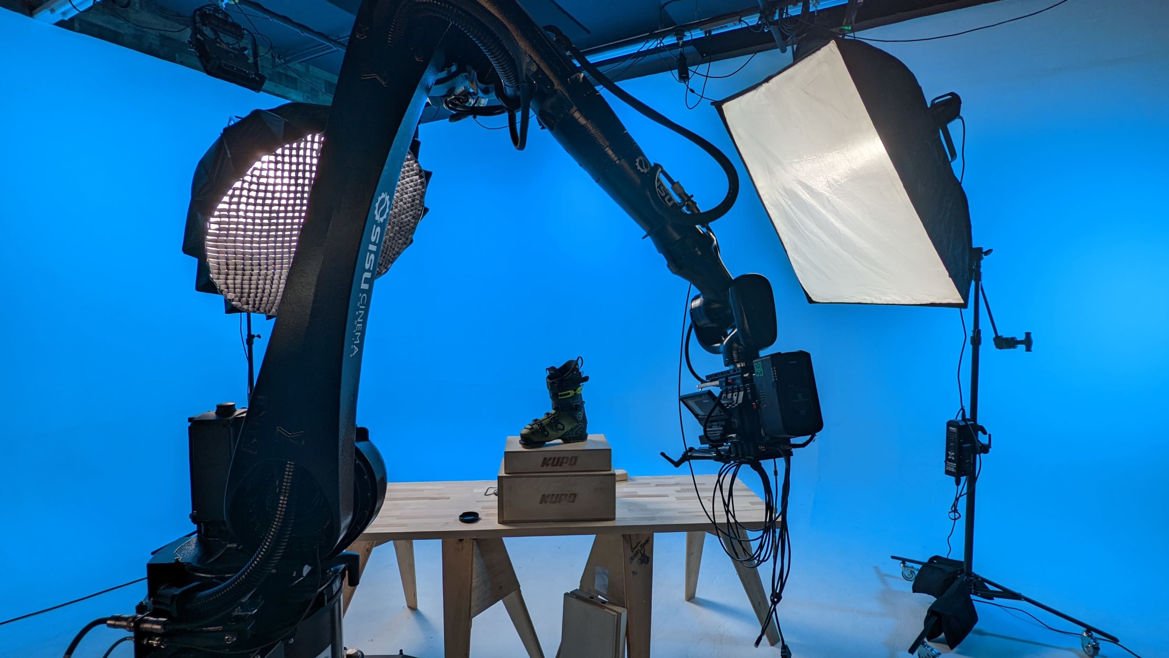 A robotic arm is positioned over a boot displayed on a pedestal, surrounded by studio lighting against a blue background.