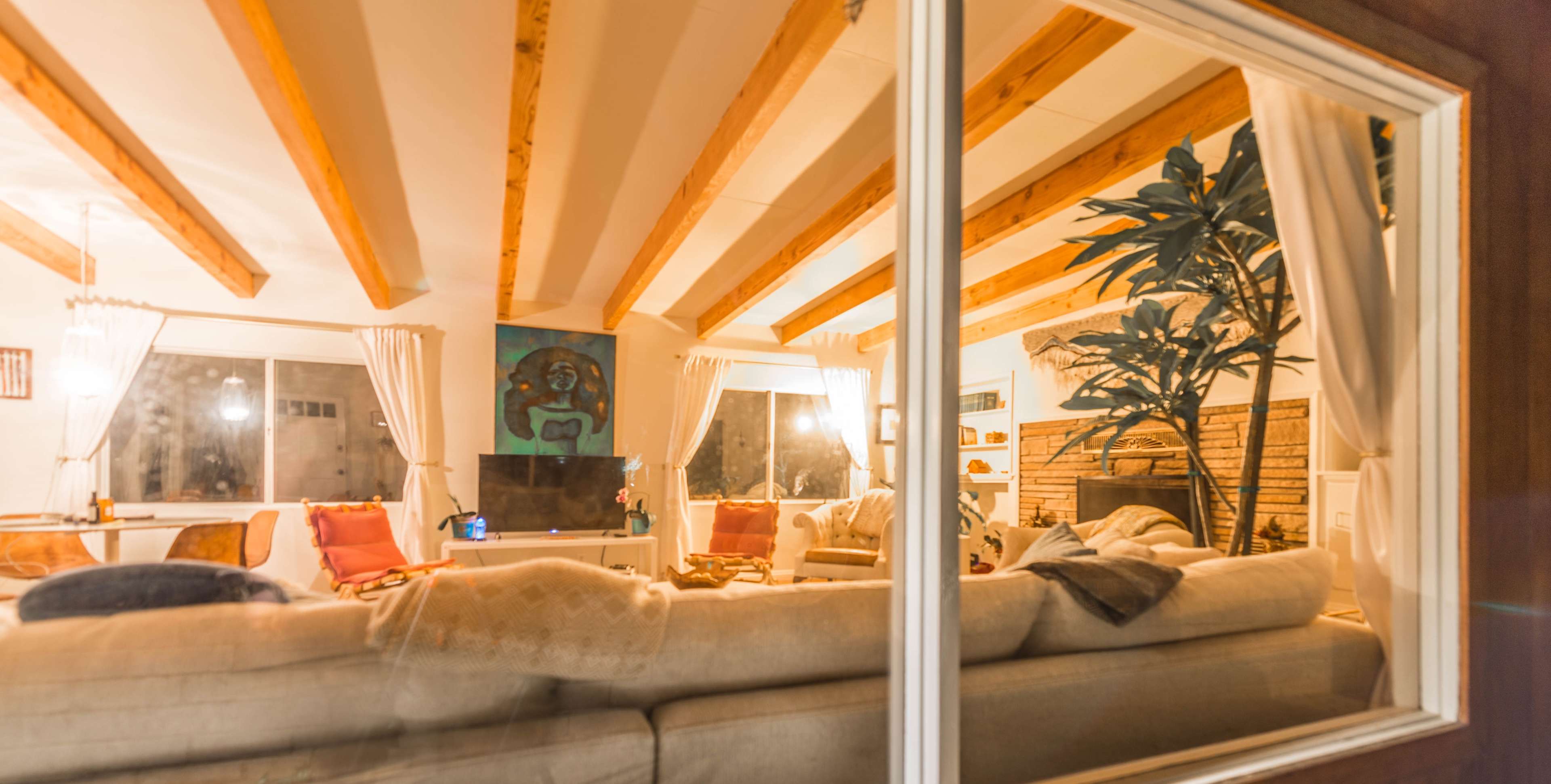 A cozy living room viewed from outside, featuring a large sectional sofa, a television, and decorative elements like a potted plant and artwork on the walls.