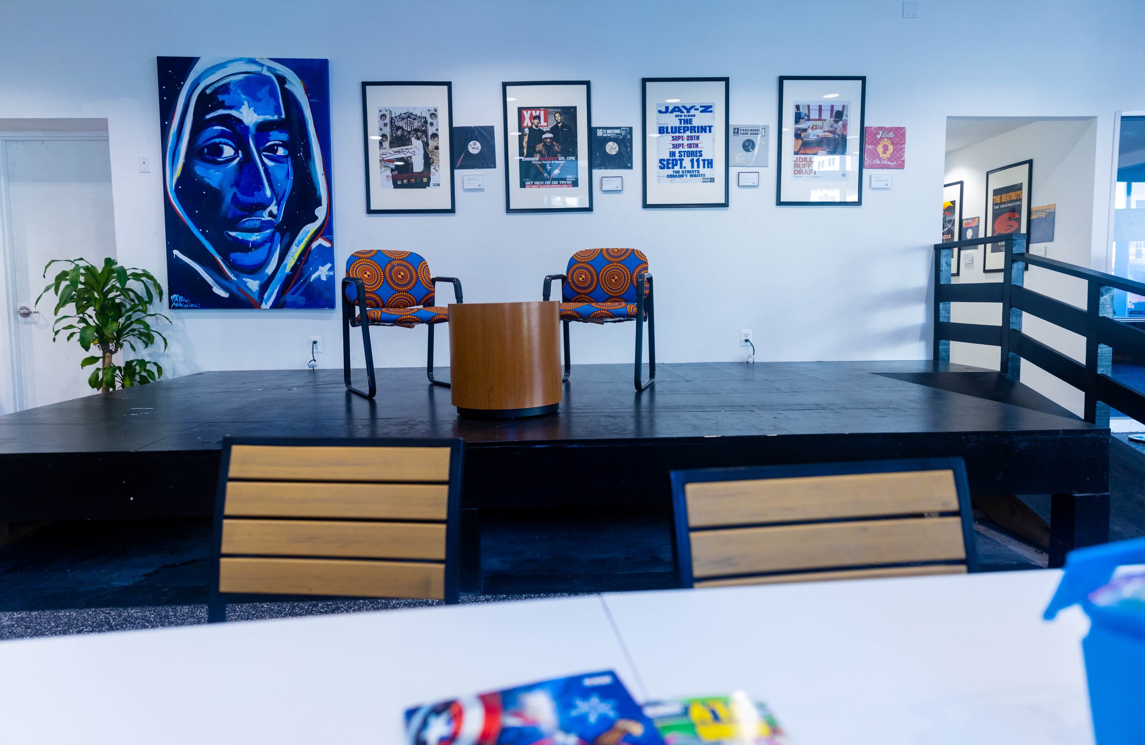 The image shows a small stage with two patterned chairs and a round table, surrounded by framed artwork and posters on the wall.