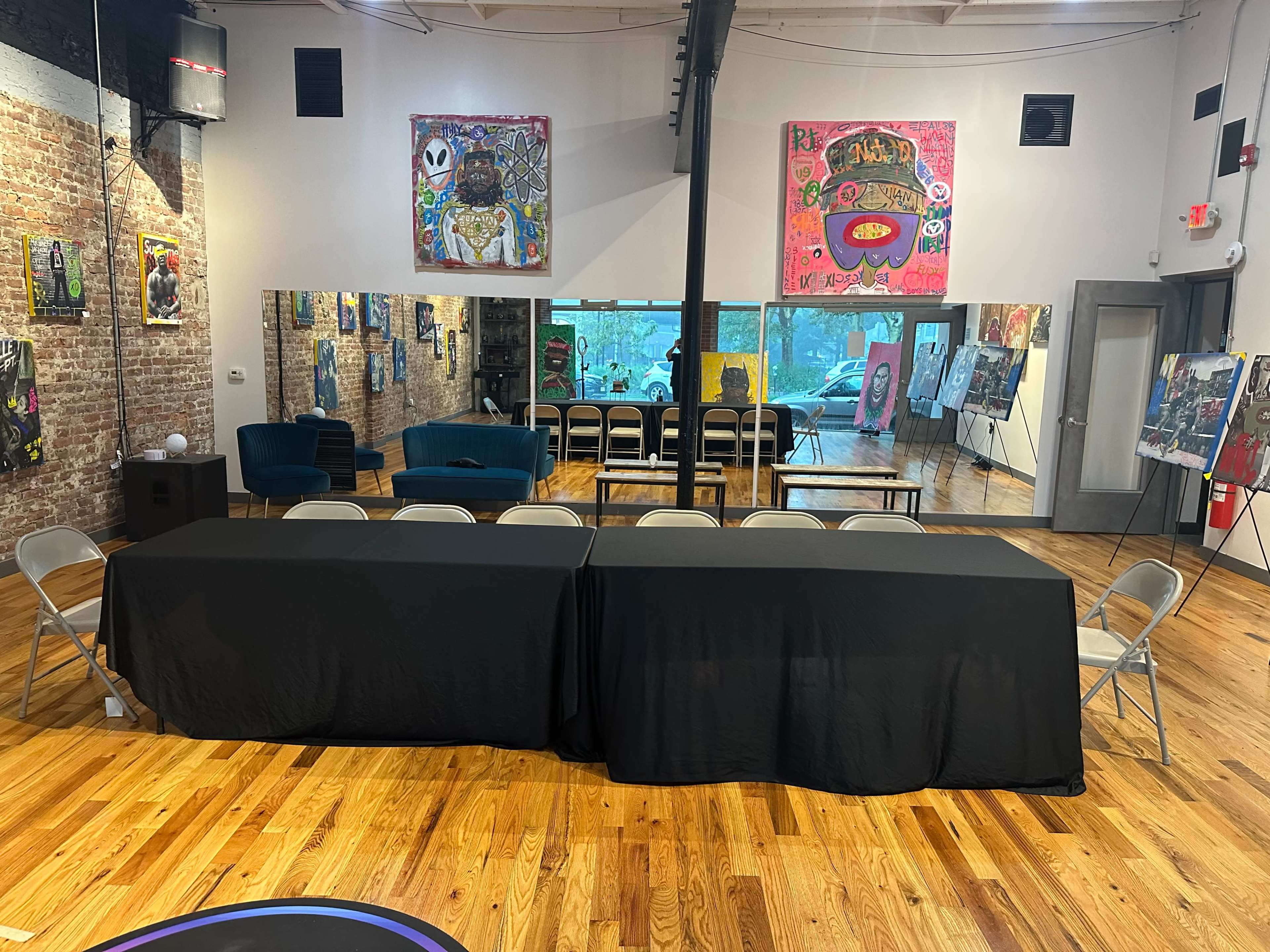 An art studio with two black tables arranged in front of a mirror, surrounded by several folding chairs, and adorned with colorful artworks on the walls.