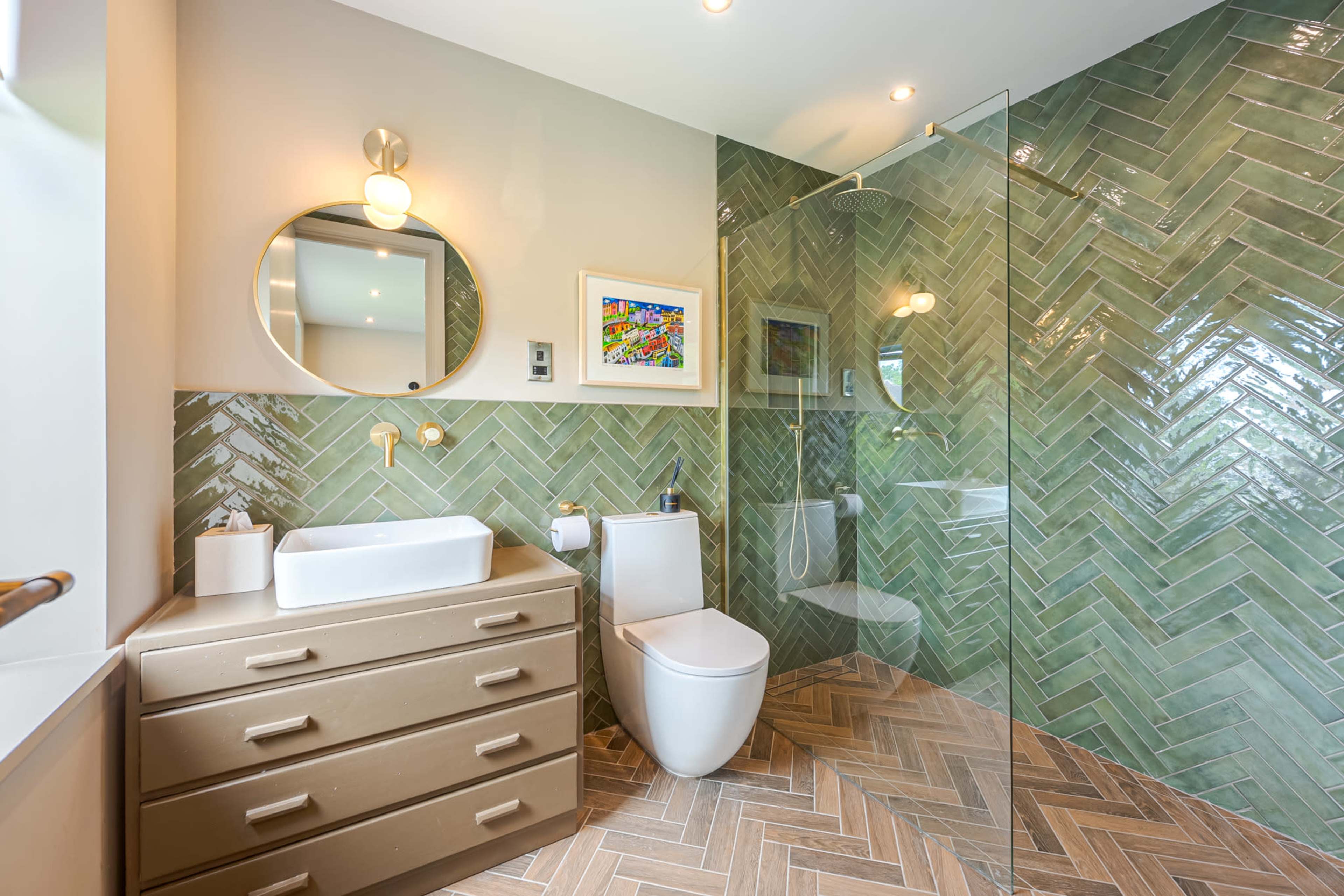 A modern bathroom featuring a herringbone-patterned floor, a vanity with a sink, a round mirror, and a glass-enclosed shower.
