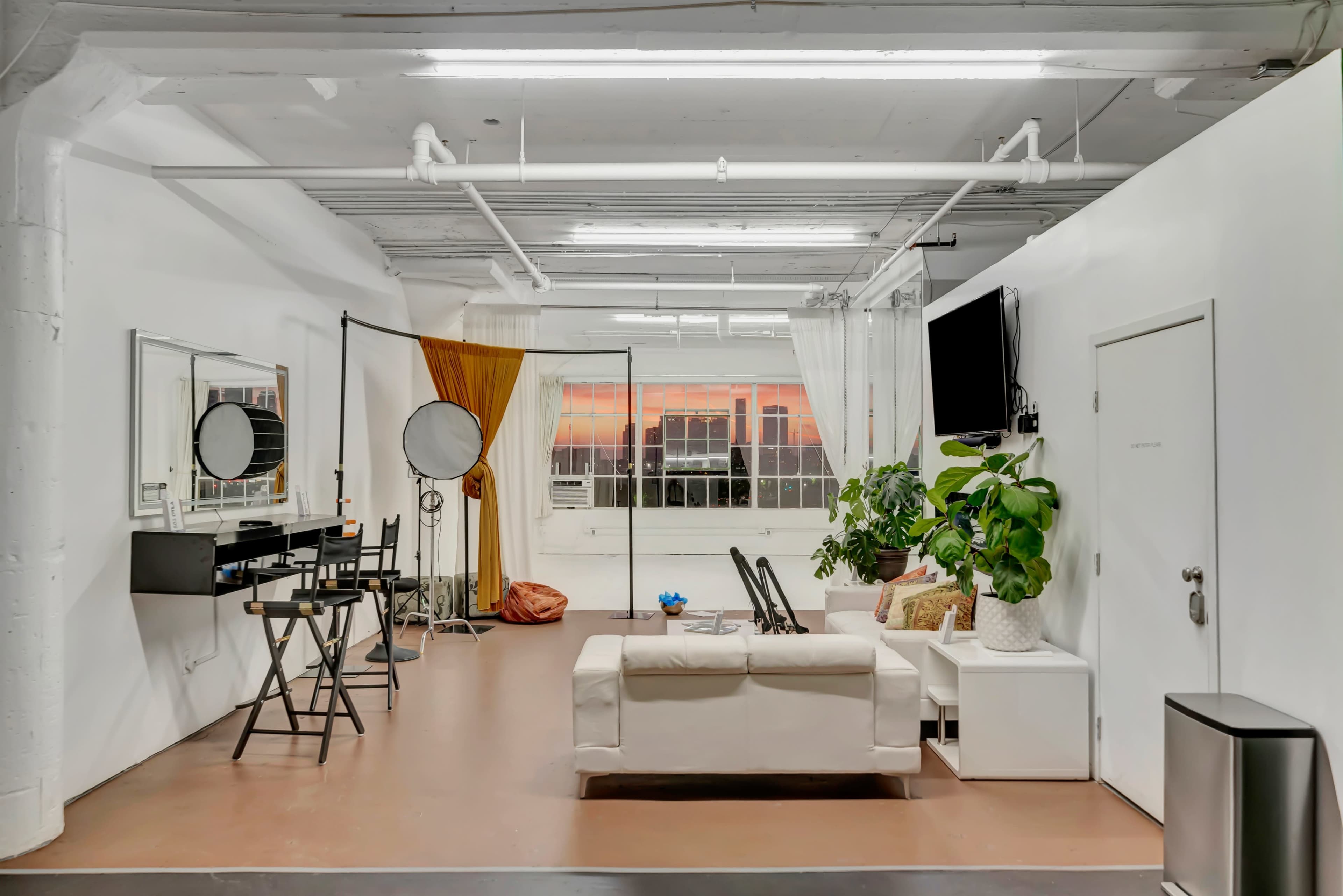 The image shows a brightly lit studio space with a white sofa, a vanity area with mirrors, and large windows revealing a sunset outside.