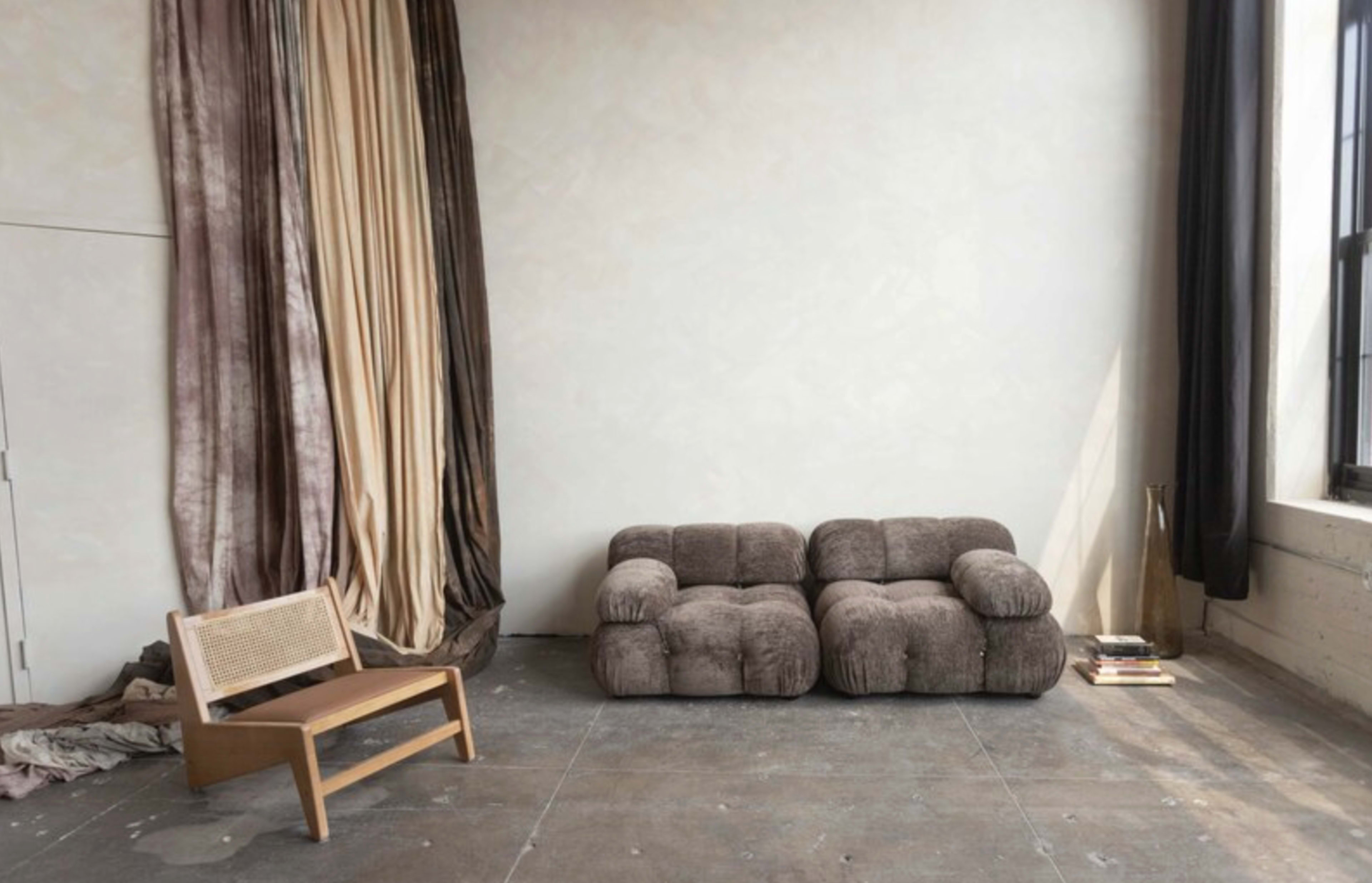 The image features a minimalist interior space with a brown cushioned sofa and a wooden chair beside textured curtains in varying colors.