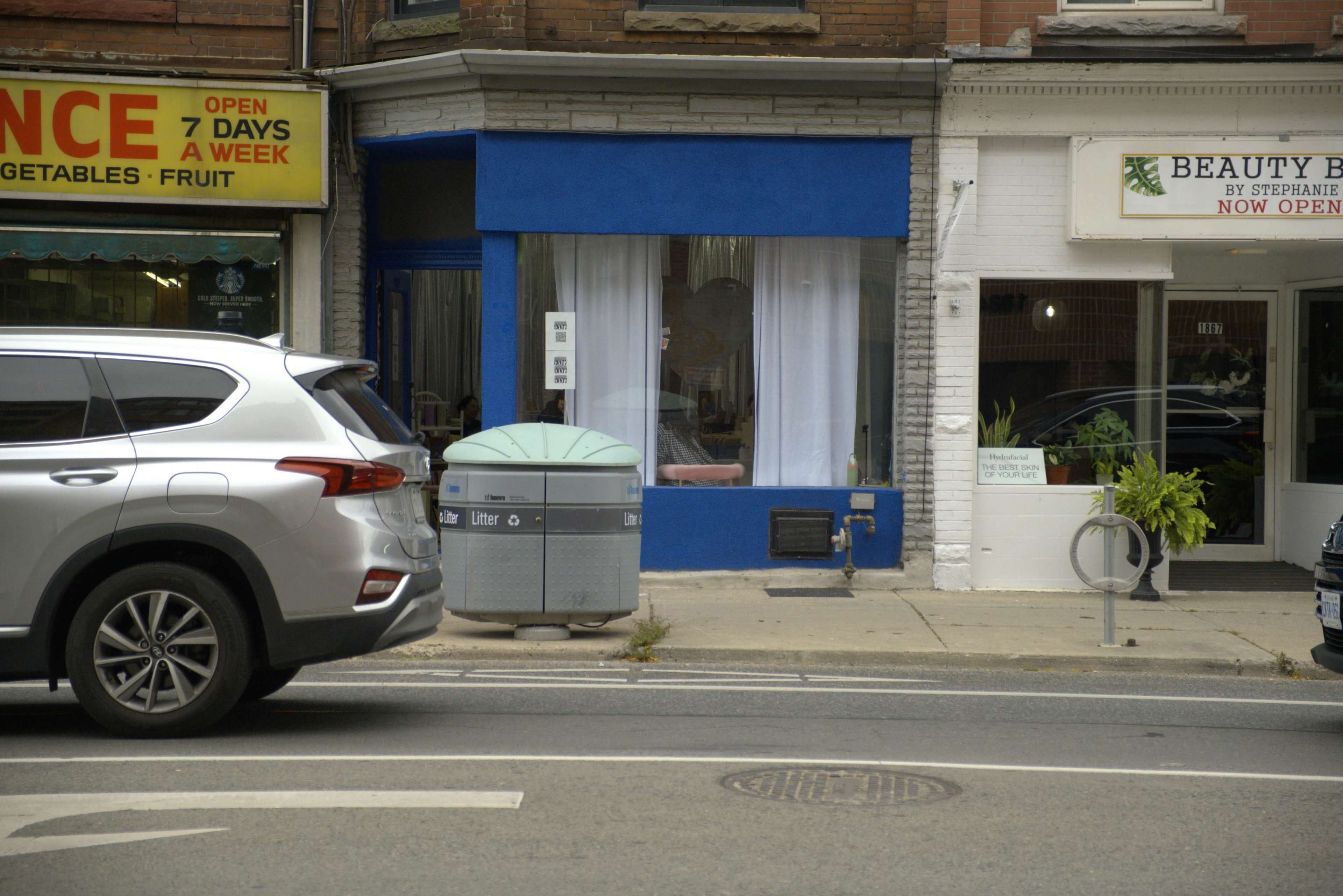 The image features a streetscape with a gray SUV parked beside a blue wall, alongside a storefront displaying a sign for a beauty salon and a grocery store.