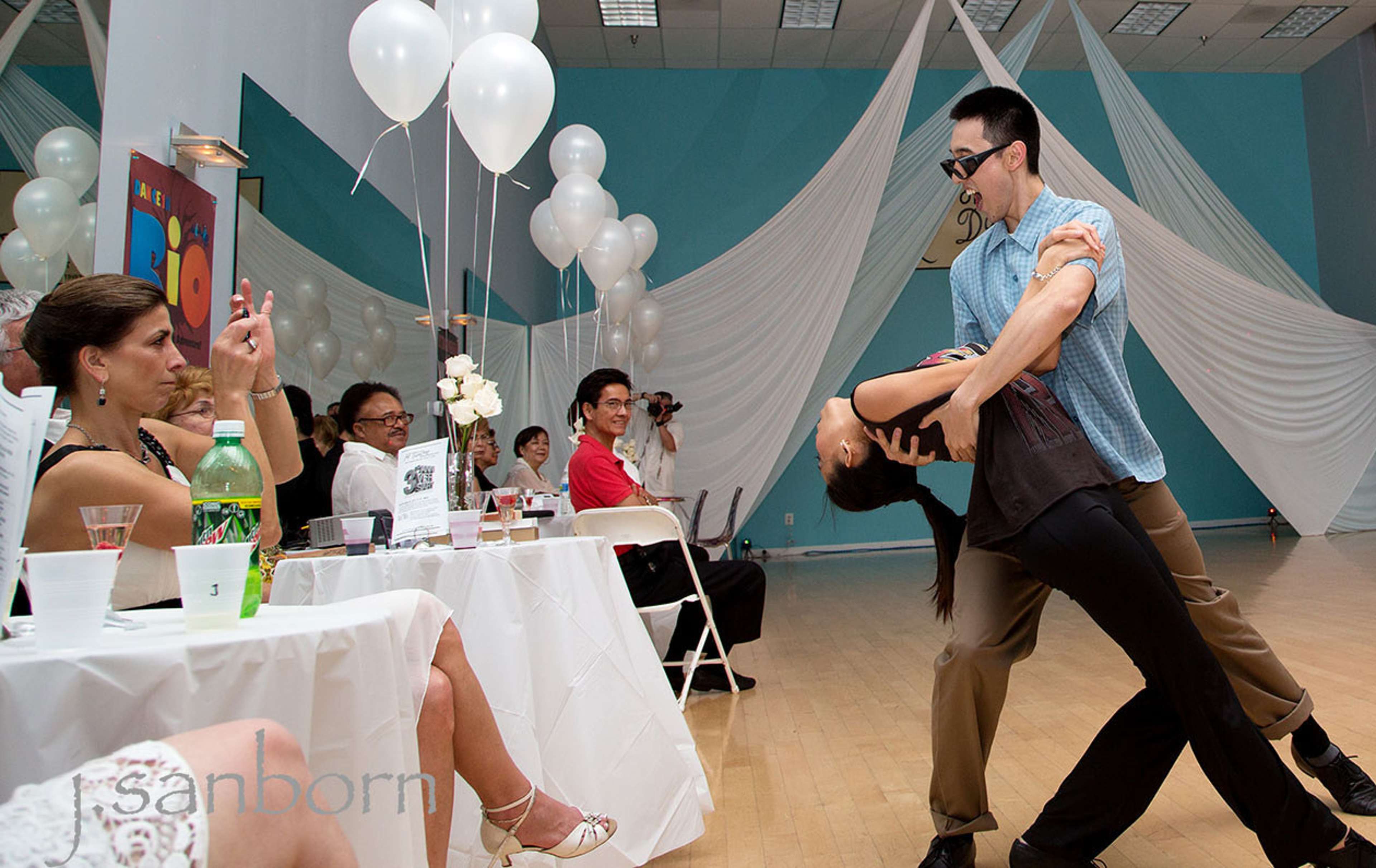 A couple performs a dance move while seated guests watch at an event decorated with balloons and table settings.