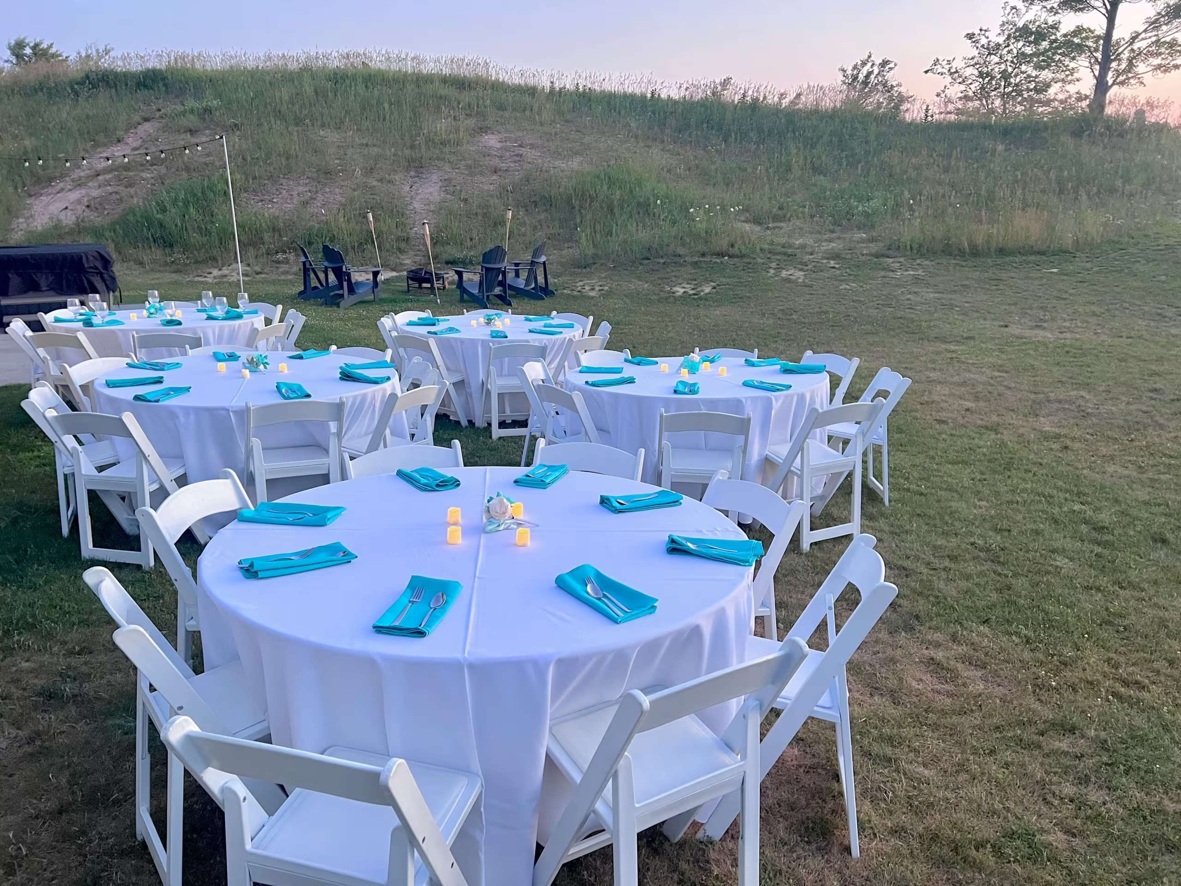 A set of round tables with white tablecloths and turquoise napkins is arranged on a grassy area near a hill, with candles in the center of each table.
