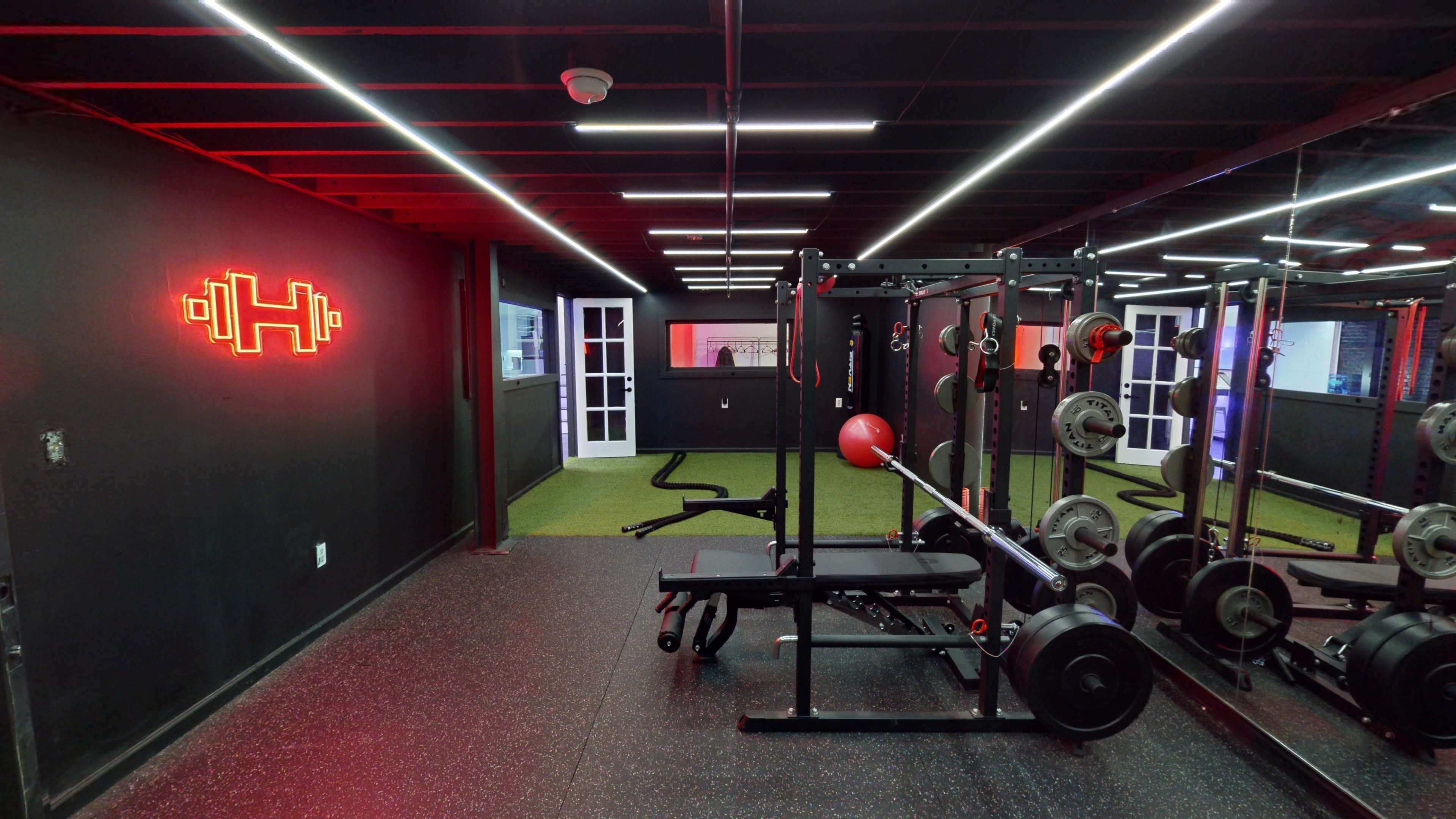 A modern gym interior featuring a weightlifting area with equipment, black rubber flooring, and neon lighting.