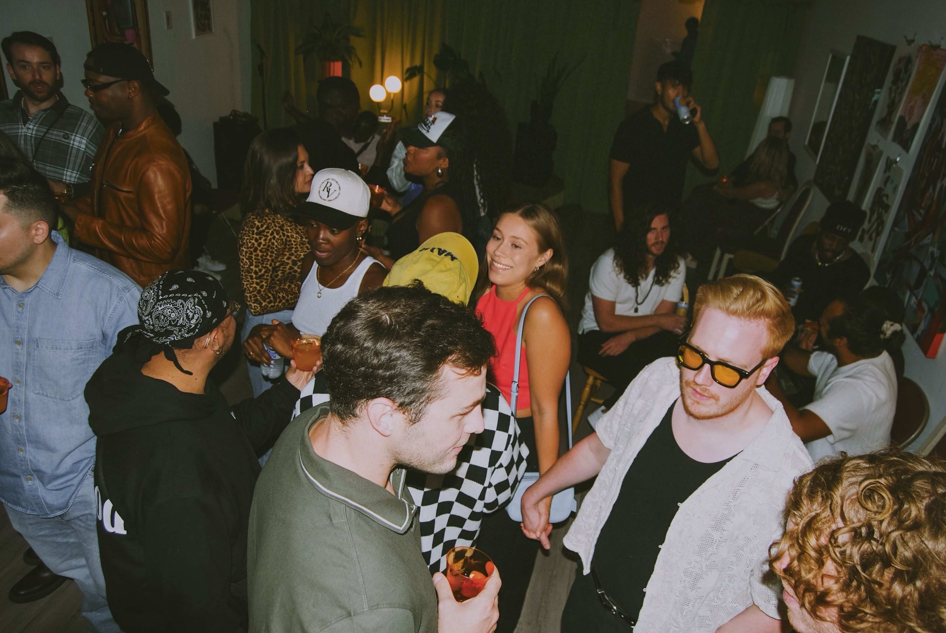 A diverse group of people socializes and dances together in a lively indoor gathering with dim lighting.