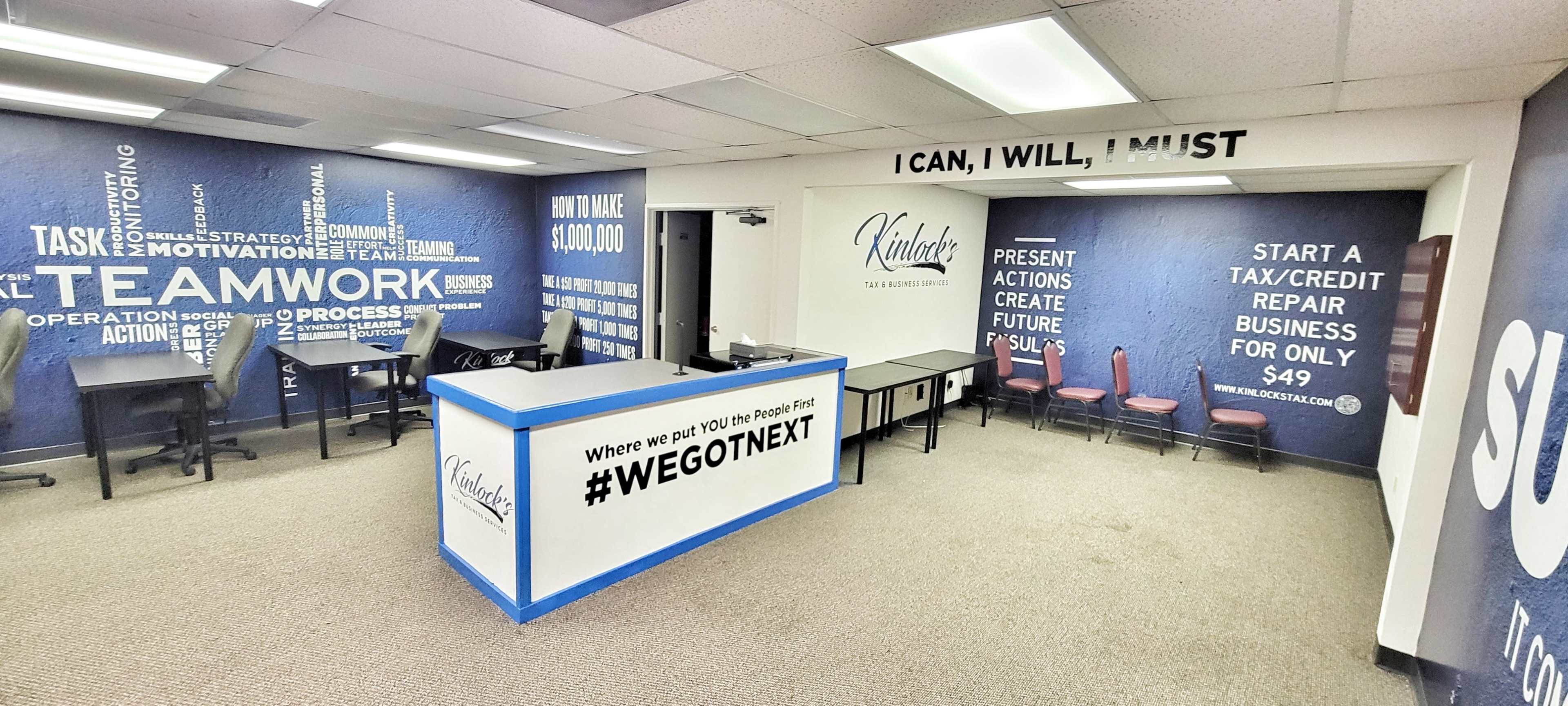 The image shows a modern office space featuring a reception area with a blue counter and motivational wall graphics, along with tables and chairs for meetings.