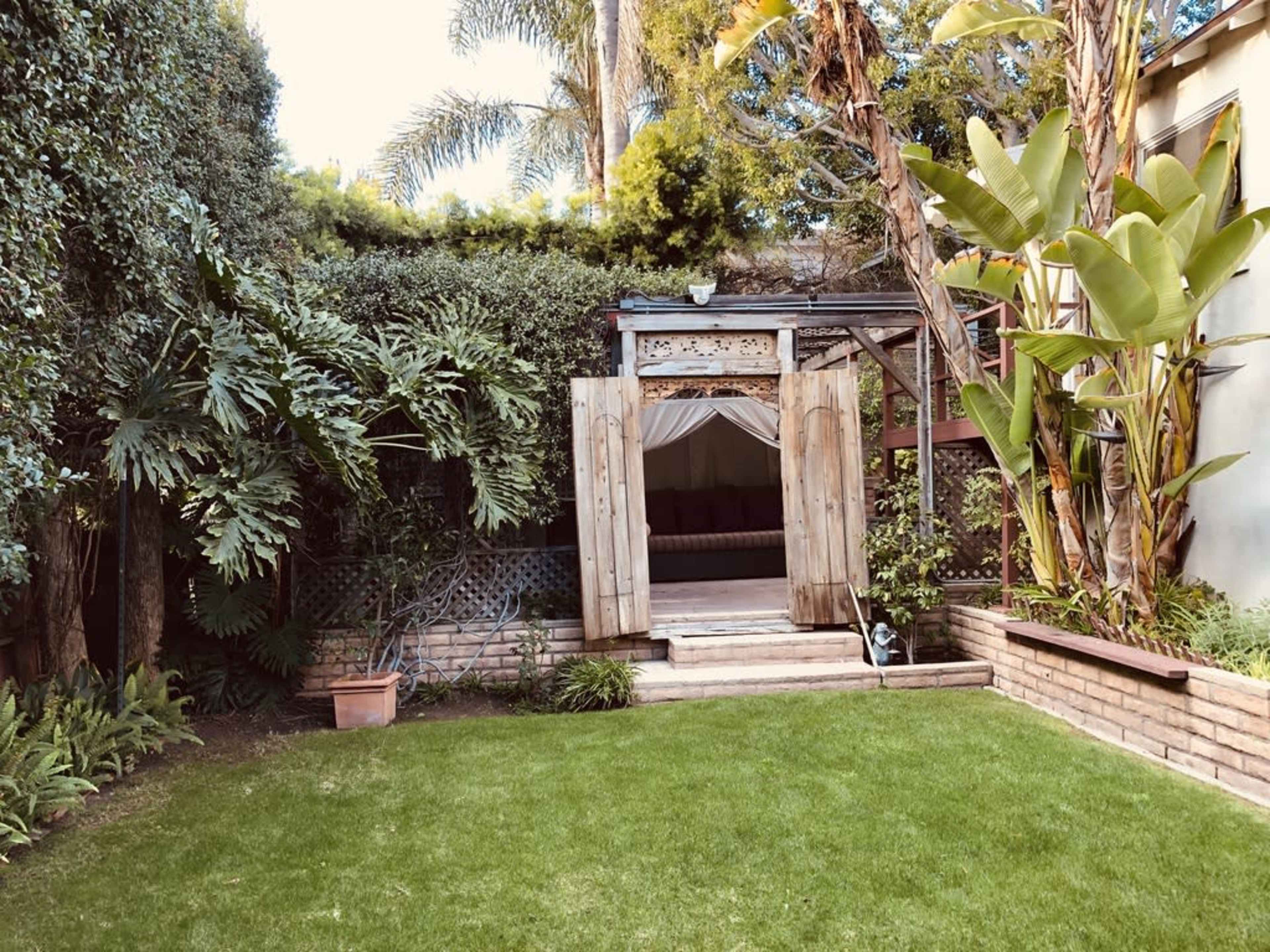 A lush garden area featuring a wooden structure with open doors, surrounded by tropical plants and a neatly maintained lawn.