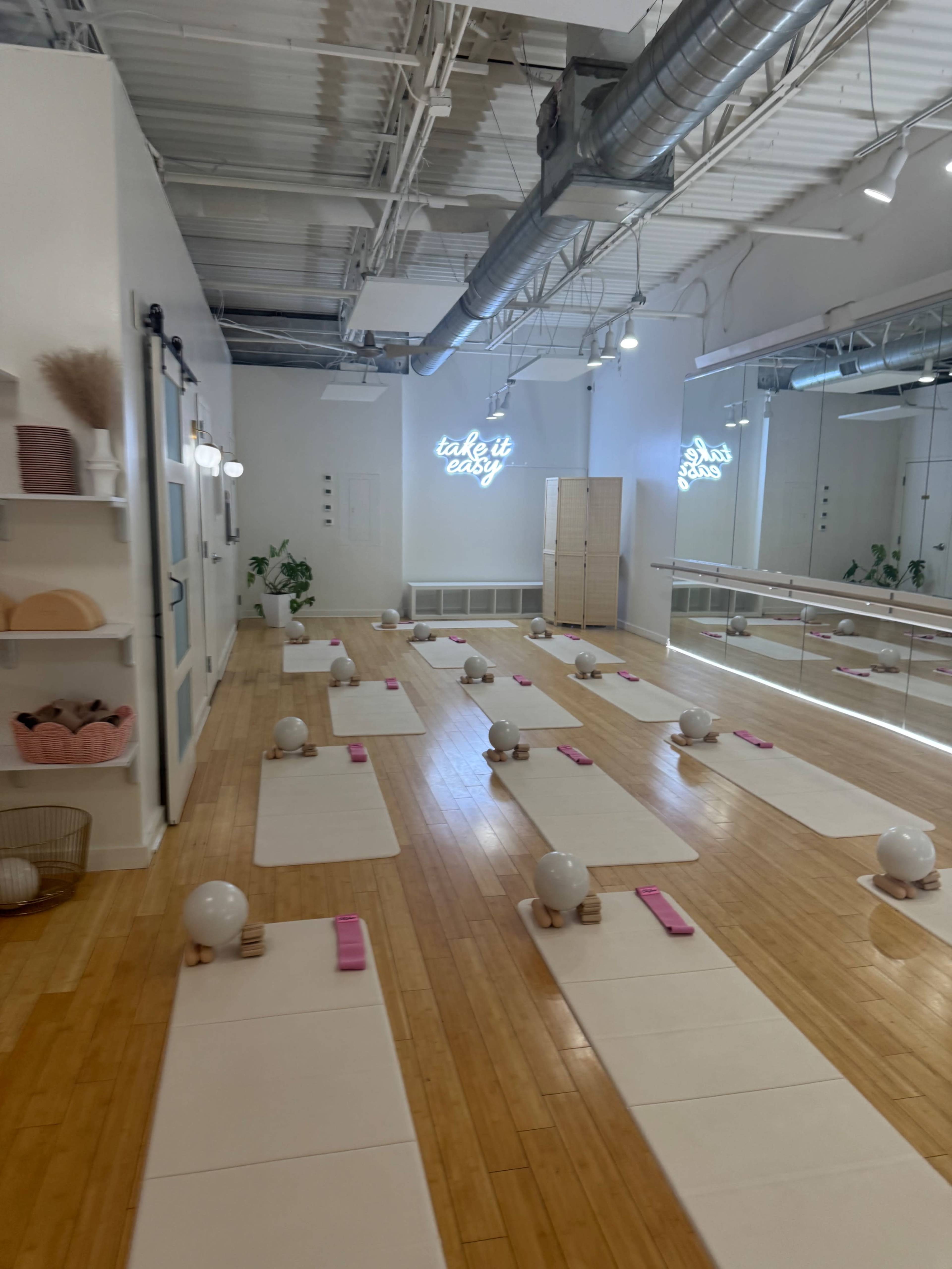 The image shows a bright, spacious yoga studio with neatly arranged mats, pink blocks, and a mirror, featuring the words "take it easy" on the wall.