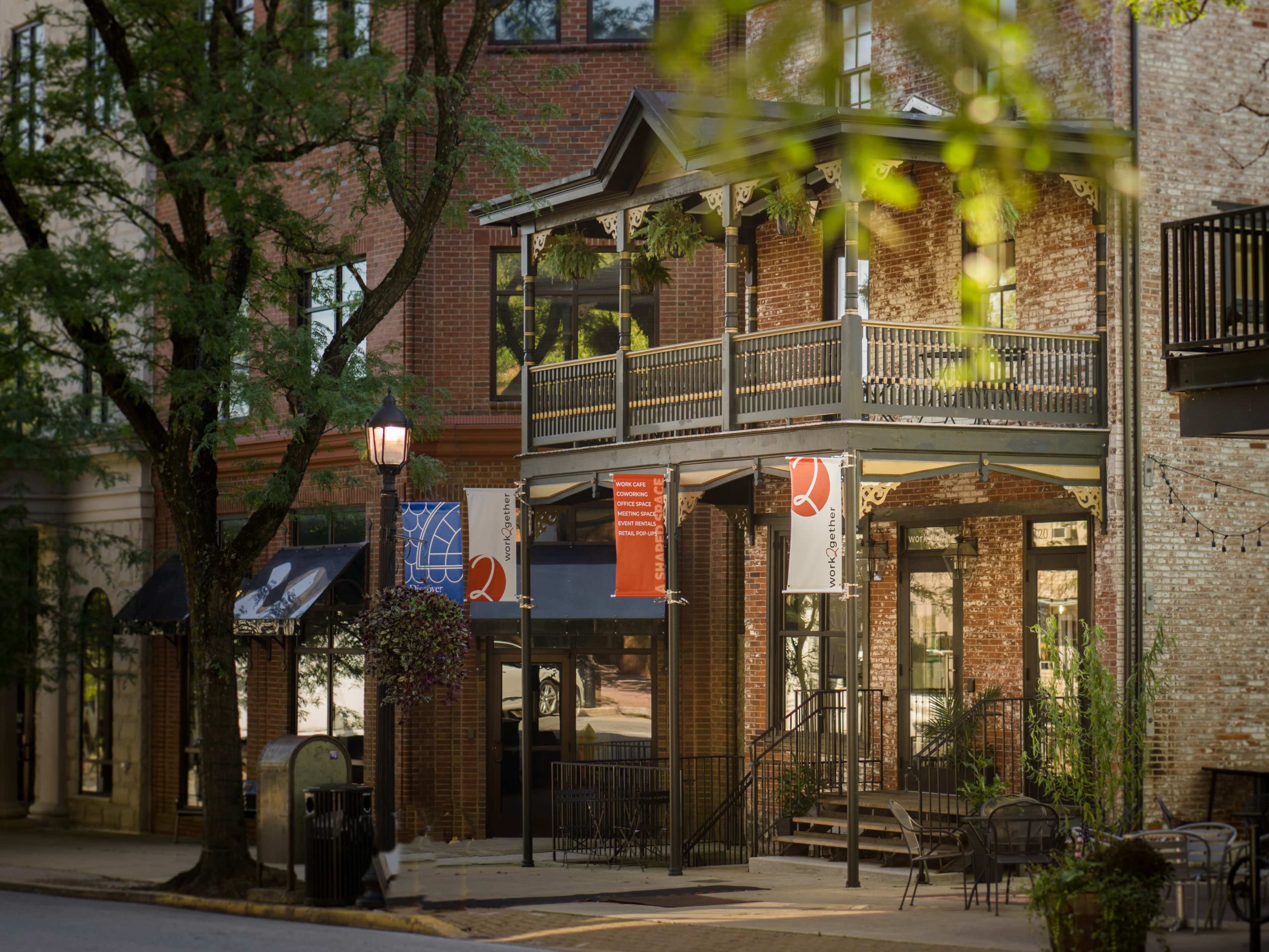 A storefront with a covered balcony, surrounded by trees and outdoor seating, in an urban setting.