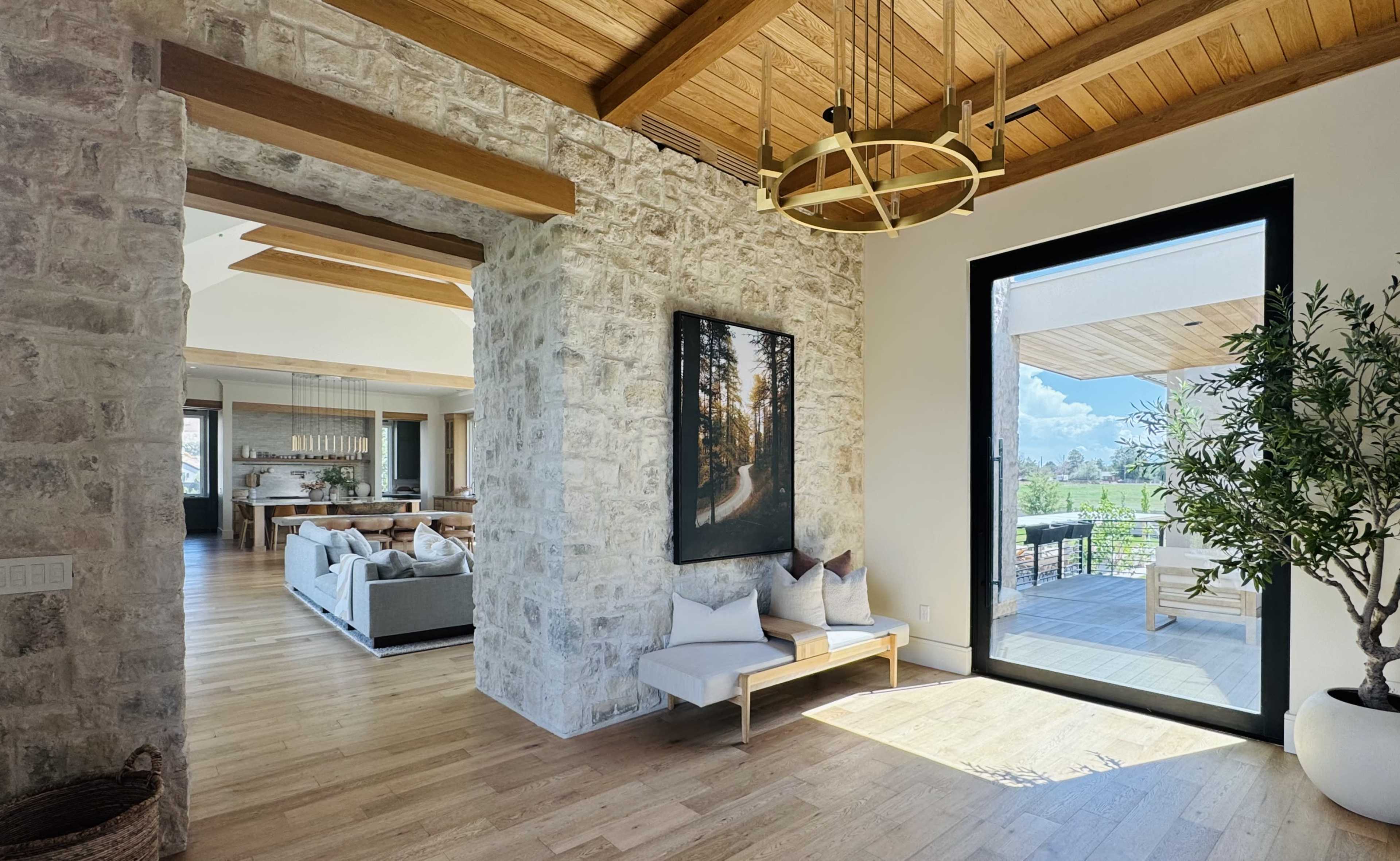 The image shows a spacious interior of a modern home with a stone wall, wooden ceiling beams, and a large glass door leading to an outdoor area.