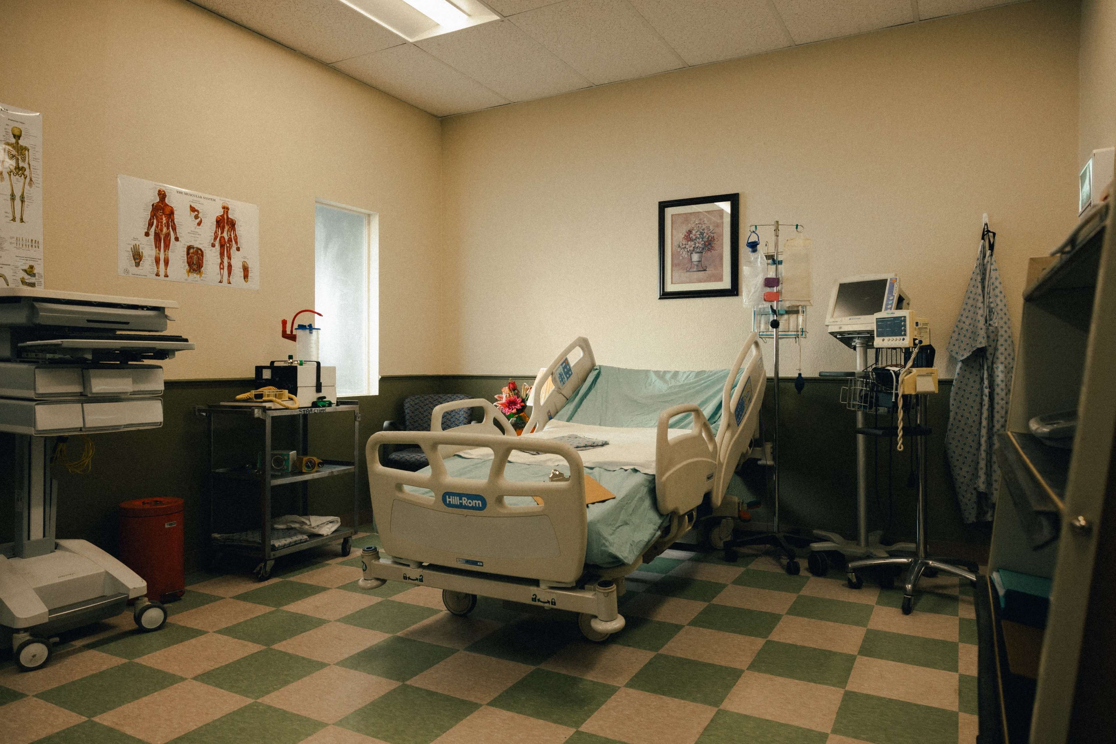 A hospital room contains a bed with blue linens, medical equipment, and anatomical posters on the wall.