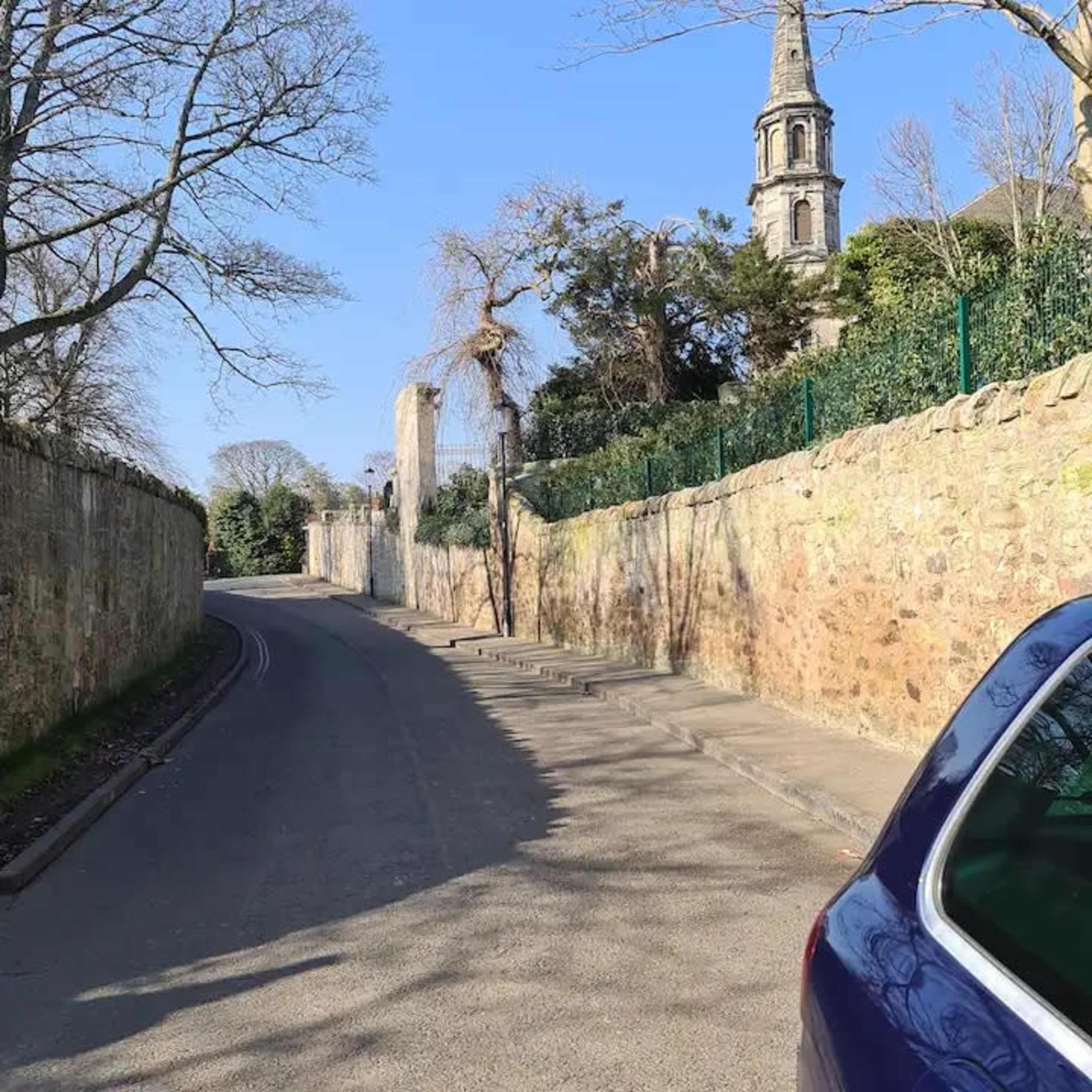 A narrow, winding road is lined with a stone wall and leads toward a church tower in the distance.