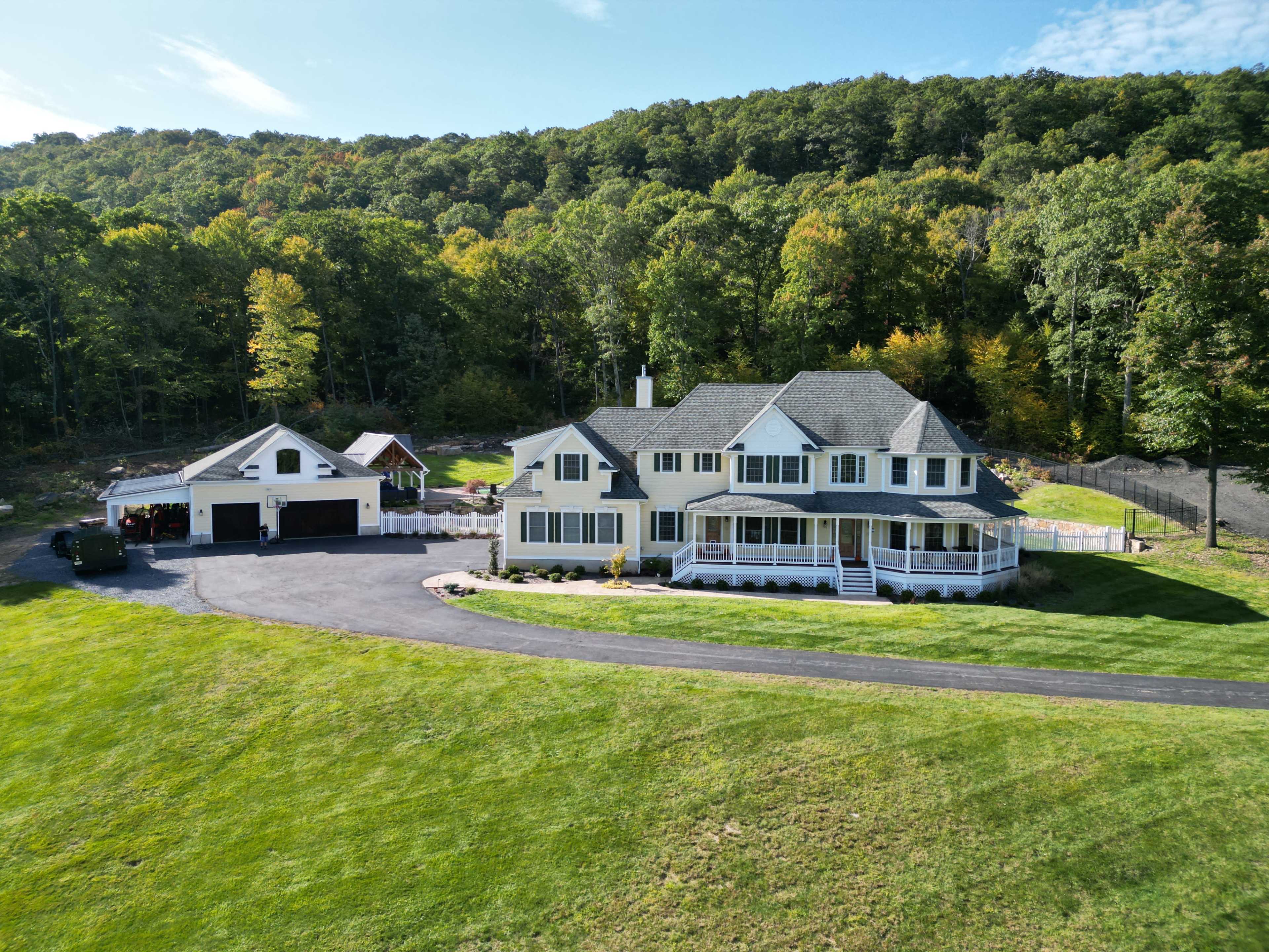 A large yellow house with a wraparound porch sits beside a garage on a landscaped property surrounded by trees.