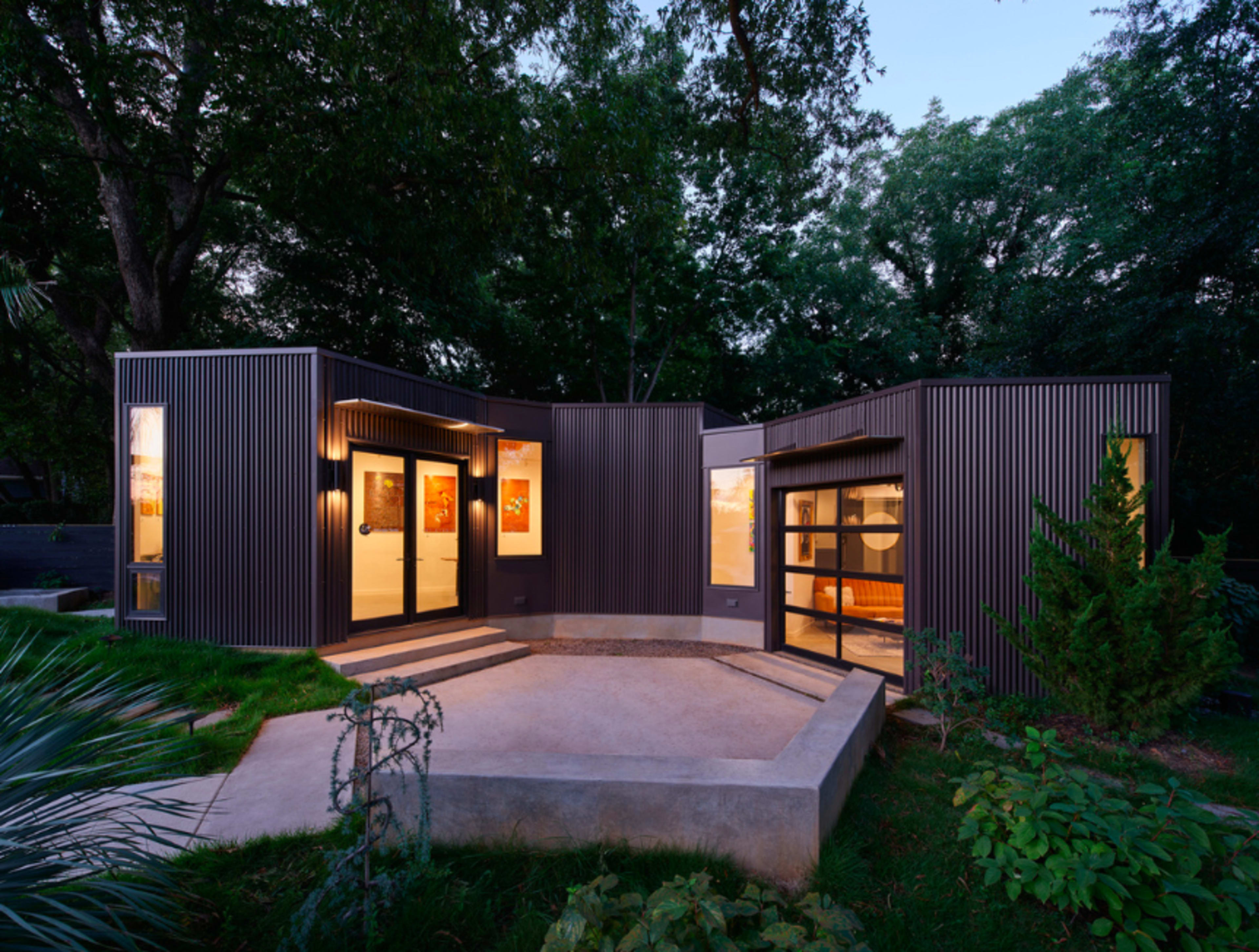 A modern, angular house with metal siding and large windows, surrounded by greenery and featuring a concrete patio.