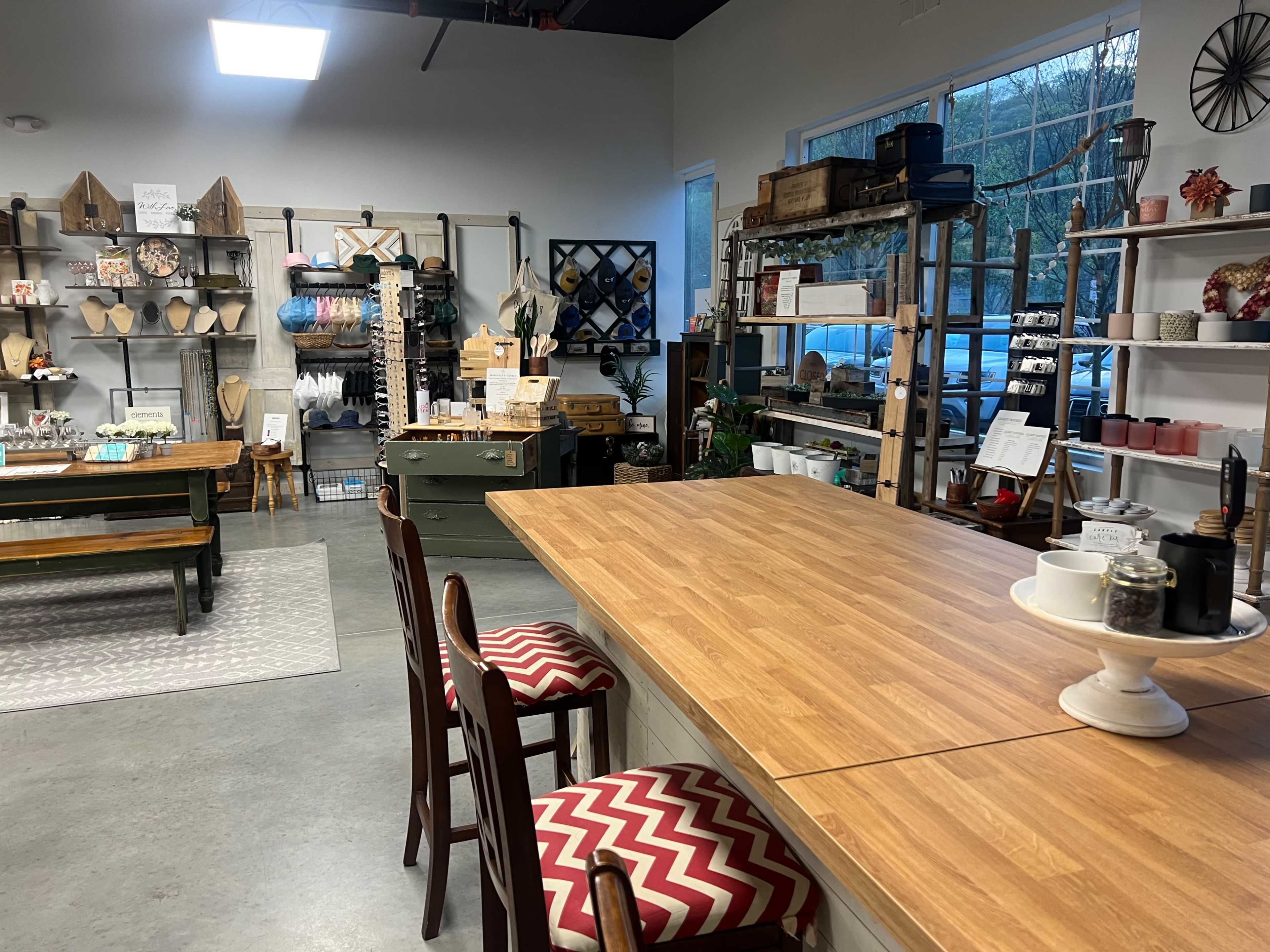 An interior space featuring wooden tables, shelves stocked with various decorative items, and a large window allowing natural light to enter.