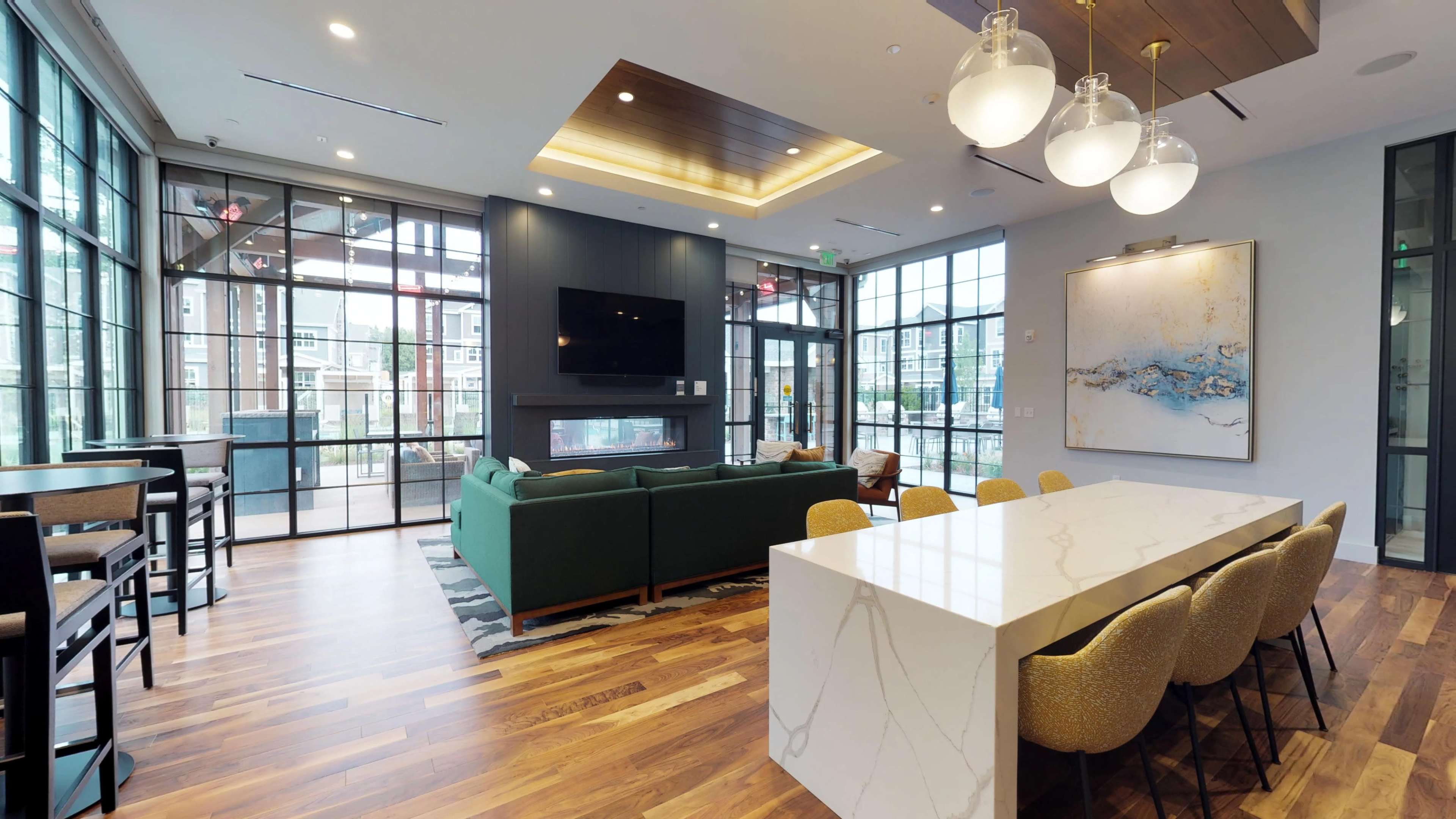 The image shows a modern living space with large windows, featuring a green sofa, a white dining table with chairs, and a wall-mounted television.