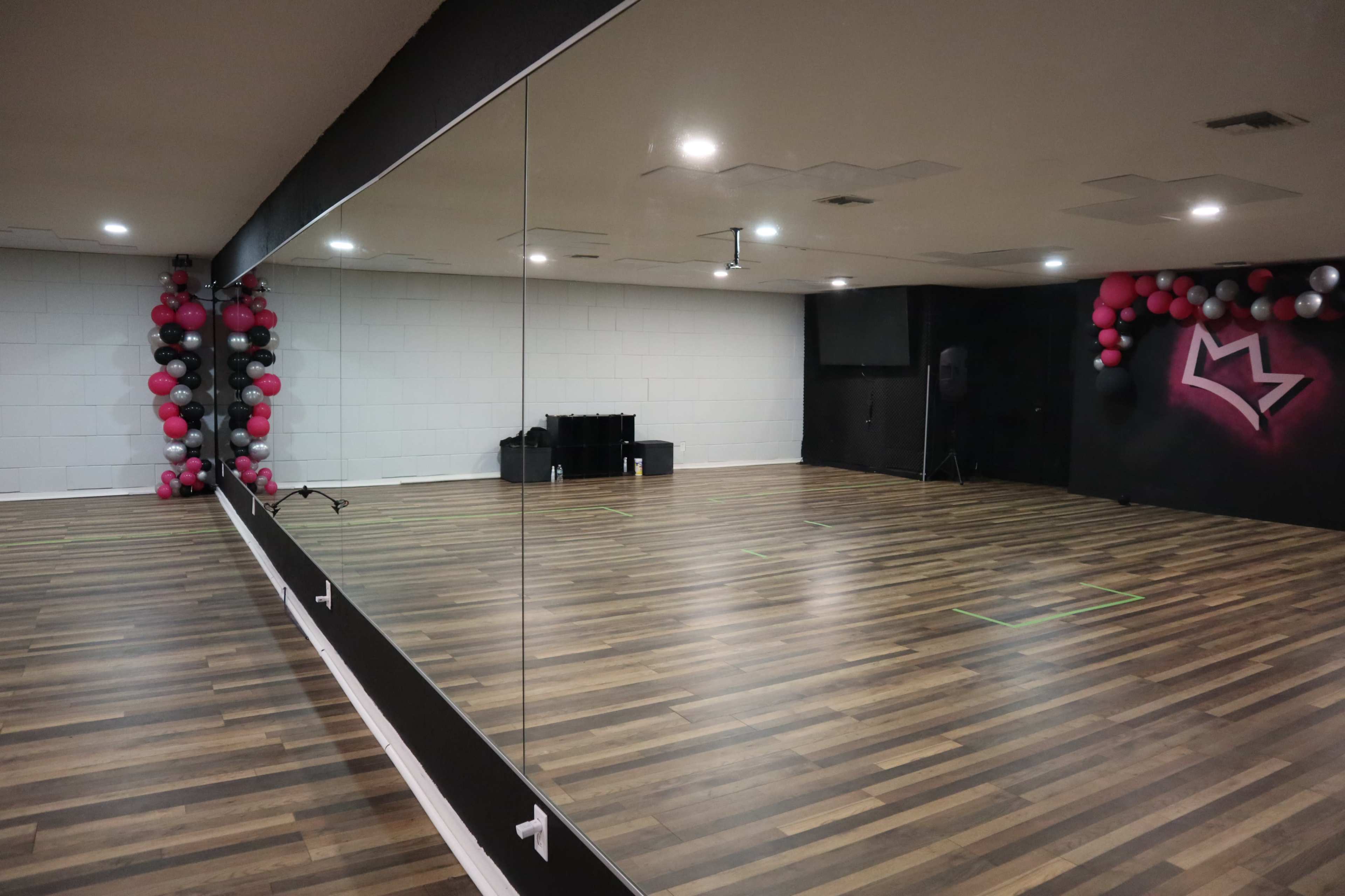 A dance studio with polished wooden floors, a large mirror on one wall, and a black wall decorated with pink and gray balloons.