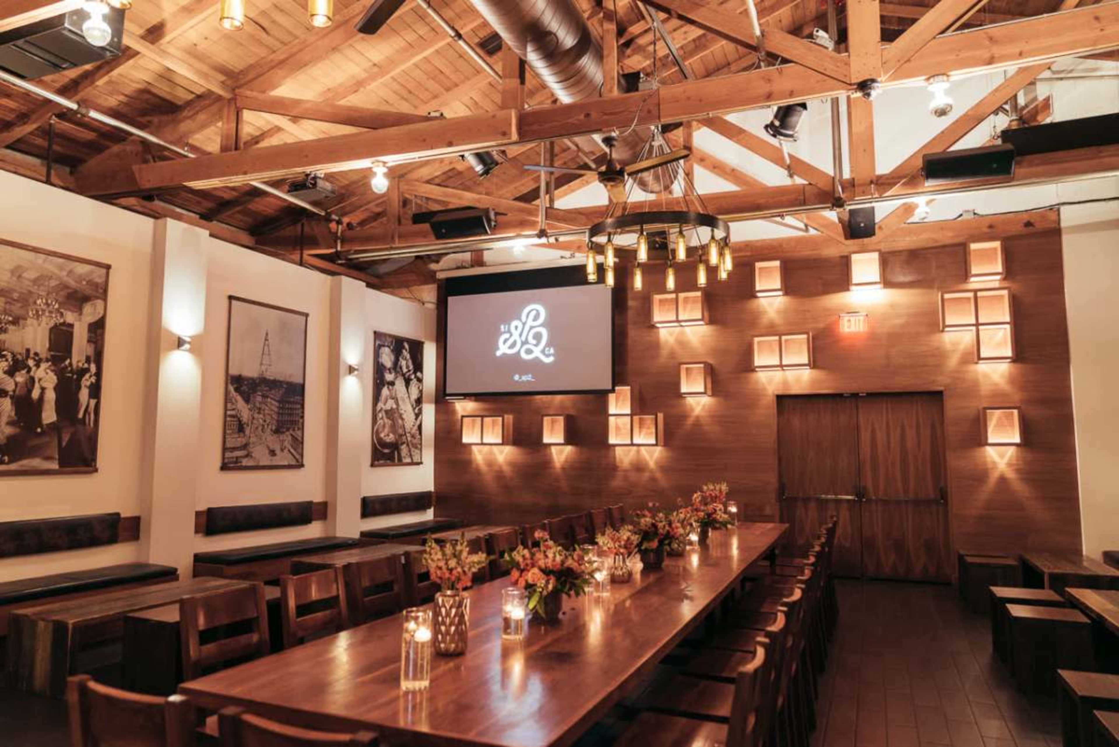 The image shows a spacious dining room with wooden beams, a long central table adorned with flowers, candles, and a projector screen displaying a logo on the wall.