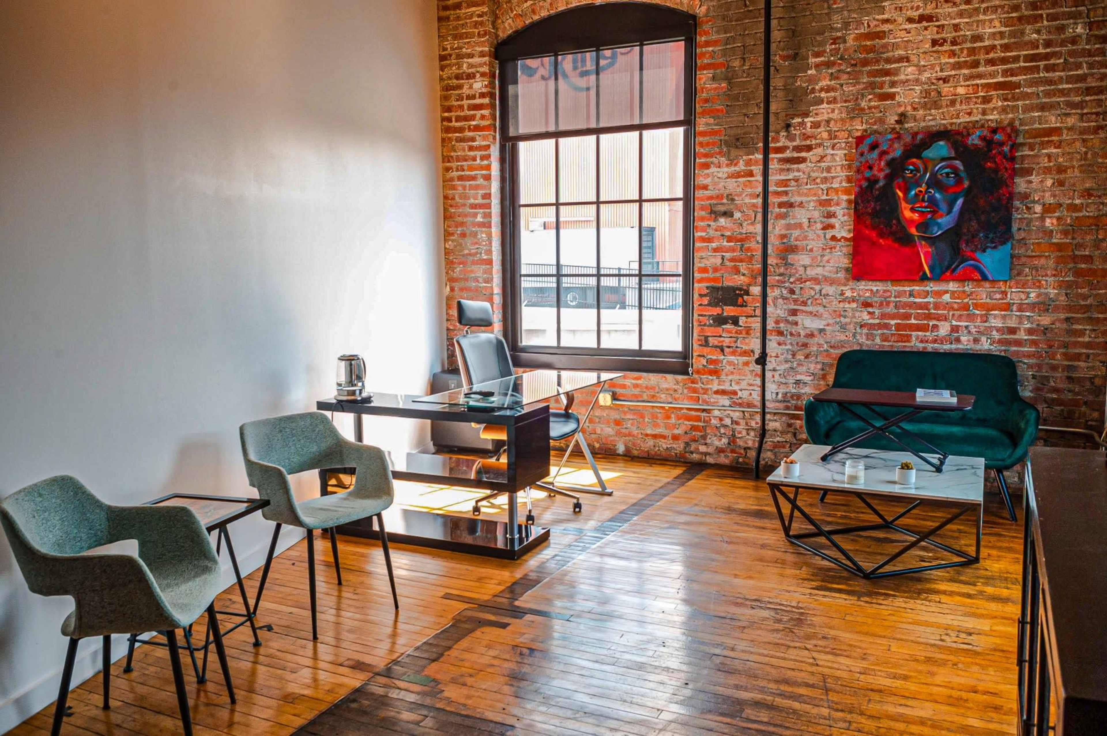 An office space featuring exposed brick walls, a wooden floor, a black desk and chair, two light gray armchairs, and a green sofa beside a coffee table.