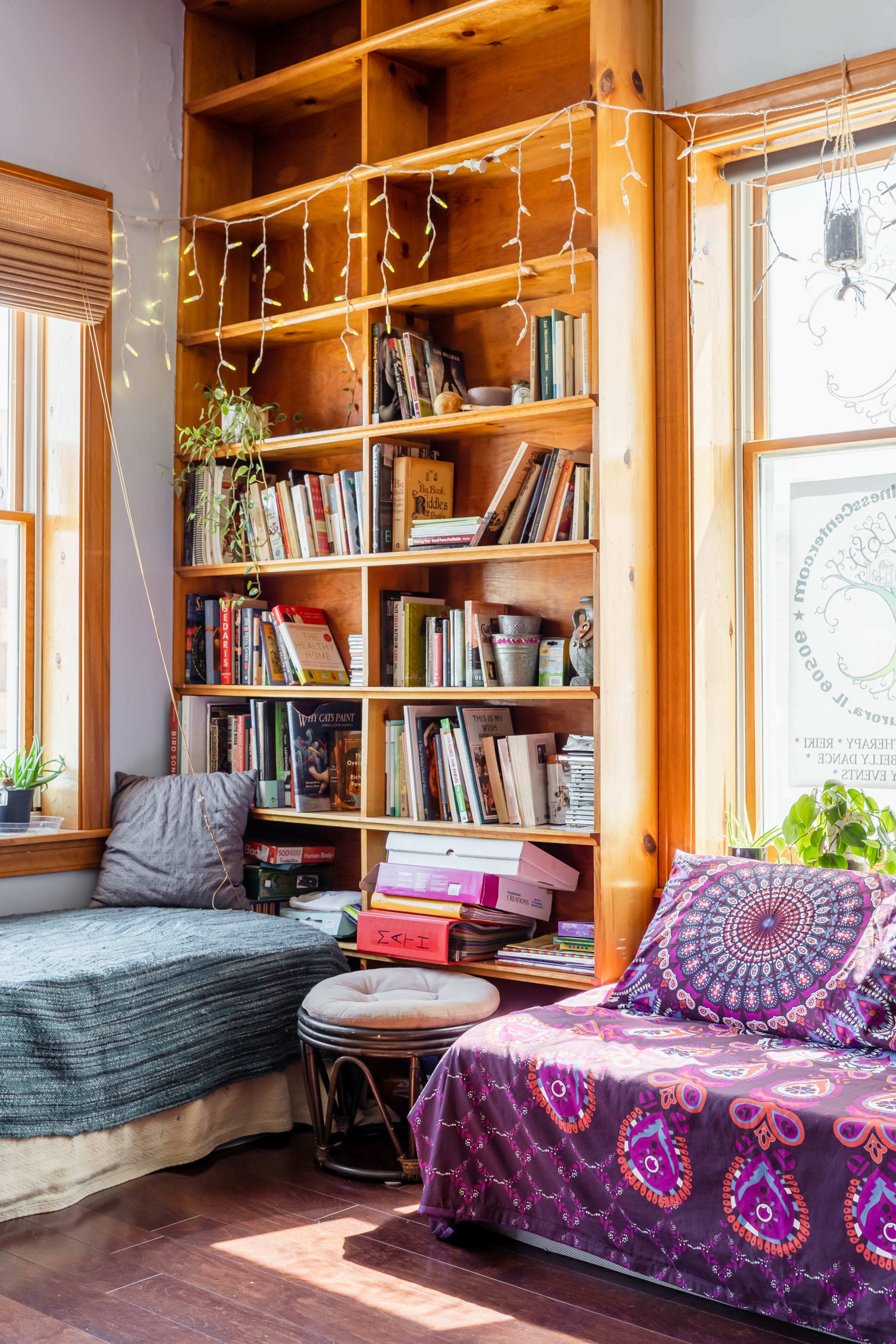 A cozy reading nook with a wooden bookshelf filled with books, a window seat adorned with cushions, and plants near large windows.