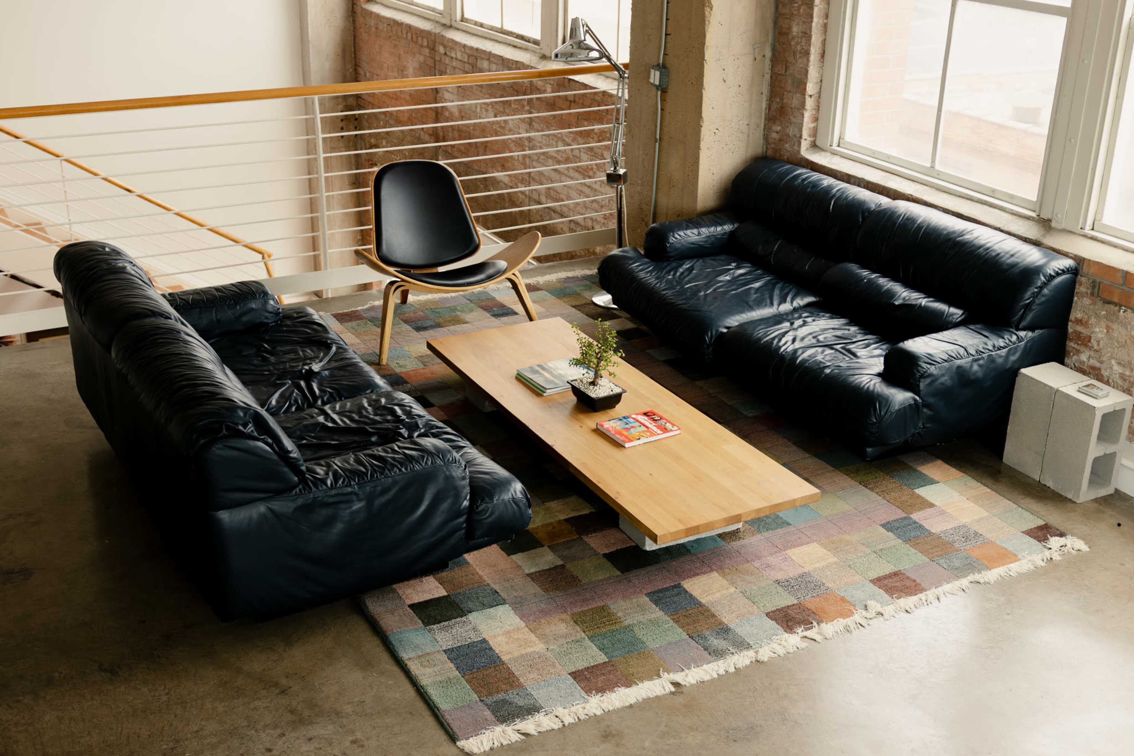 The image shows a modern living space with a coffee table, two dark leather sofas, an armchair, and a patterned area rug on a concrete floor.