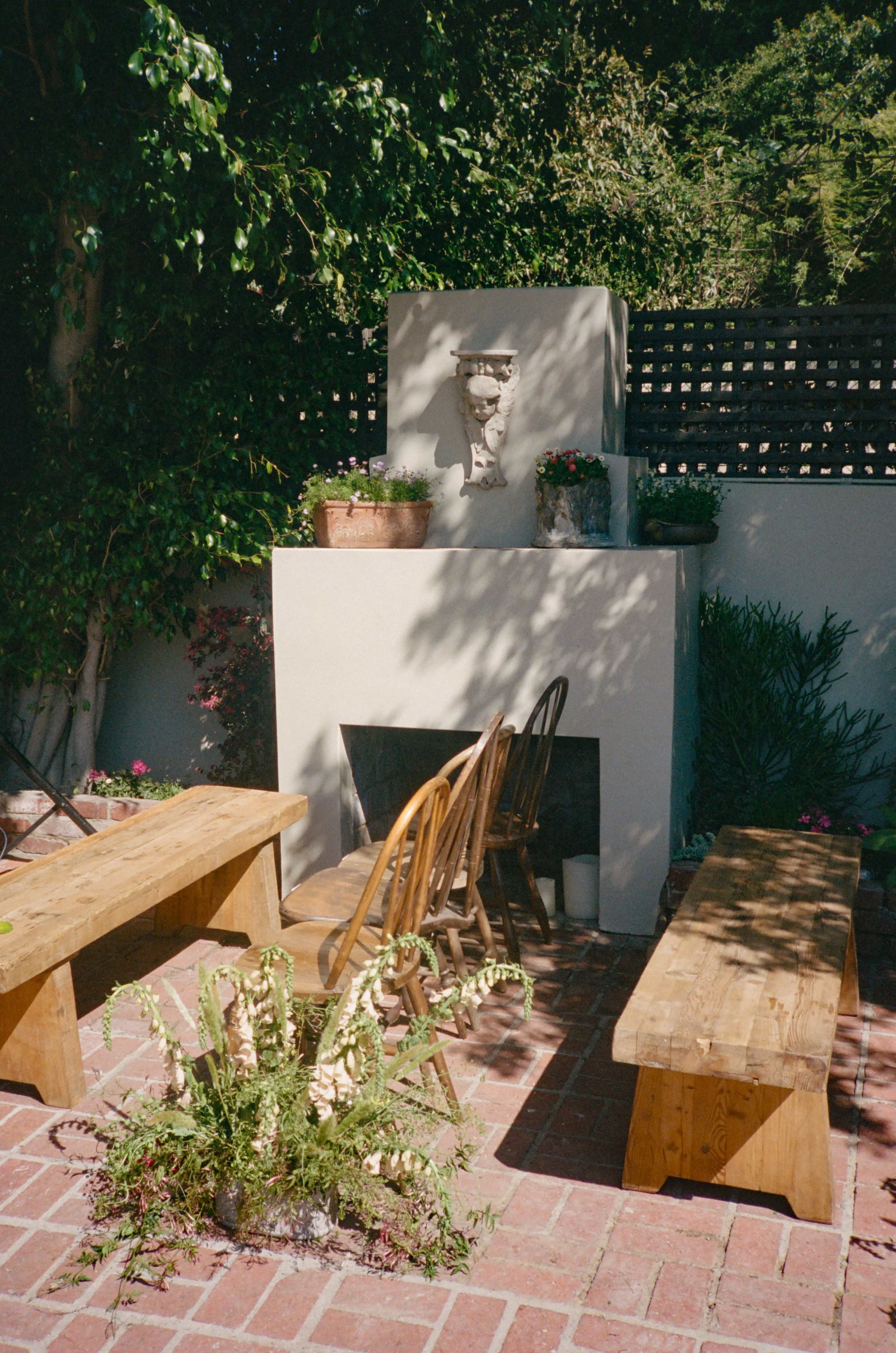 A garden patio features wooden benches, a fireplace with a bust above it, and potted plants around a brick floor.