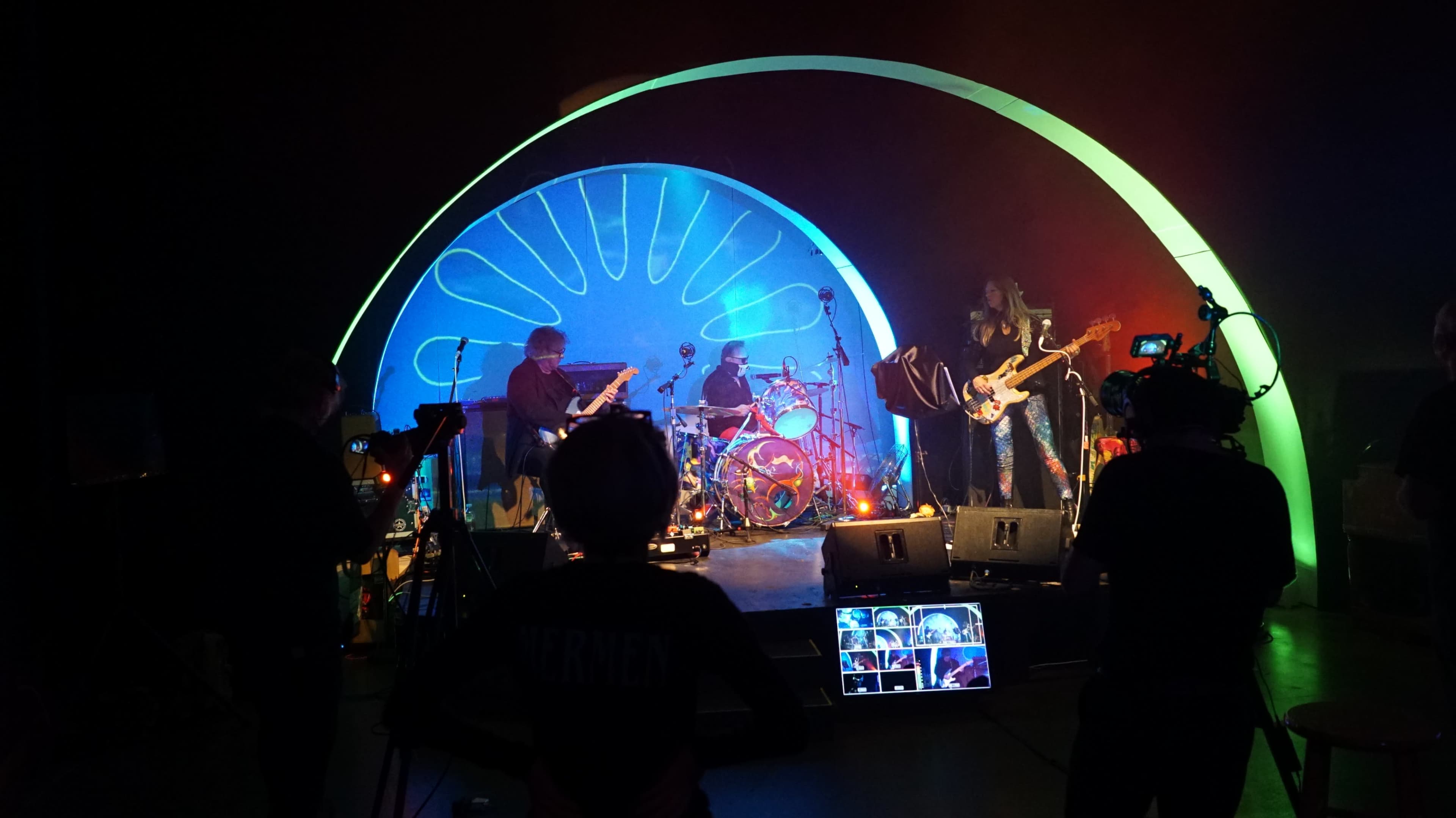 A band performs on stage with colorful lighting and a large backdrop, while cameras capture the scene.