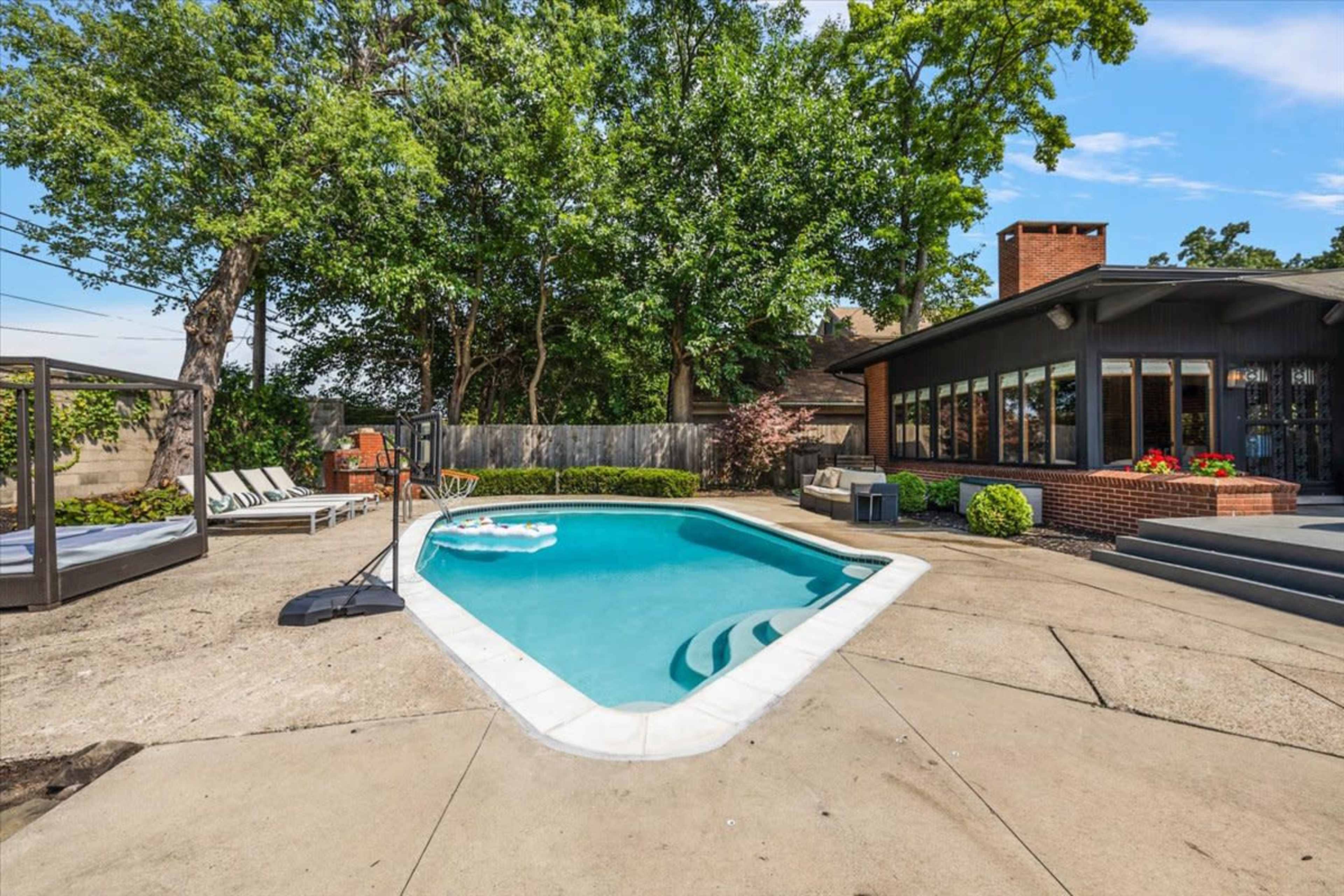 The image shows a rectangular swimming pool surrounded by concrete, lounge chairs, and a shaded seating area, with trees and a house in the background.