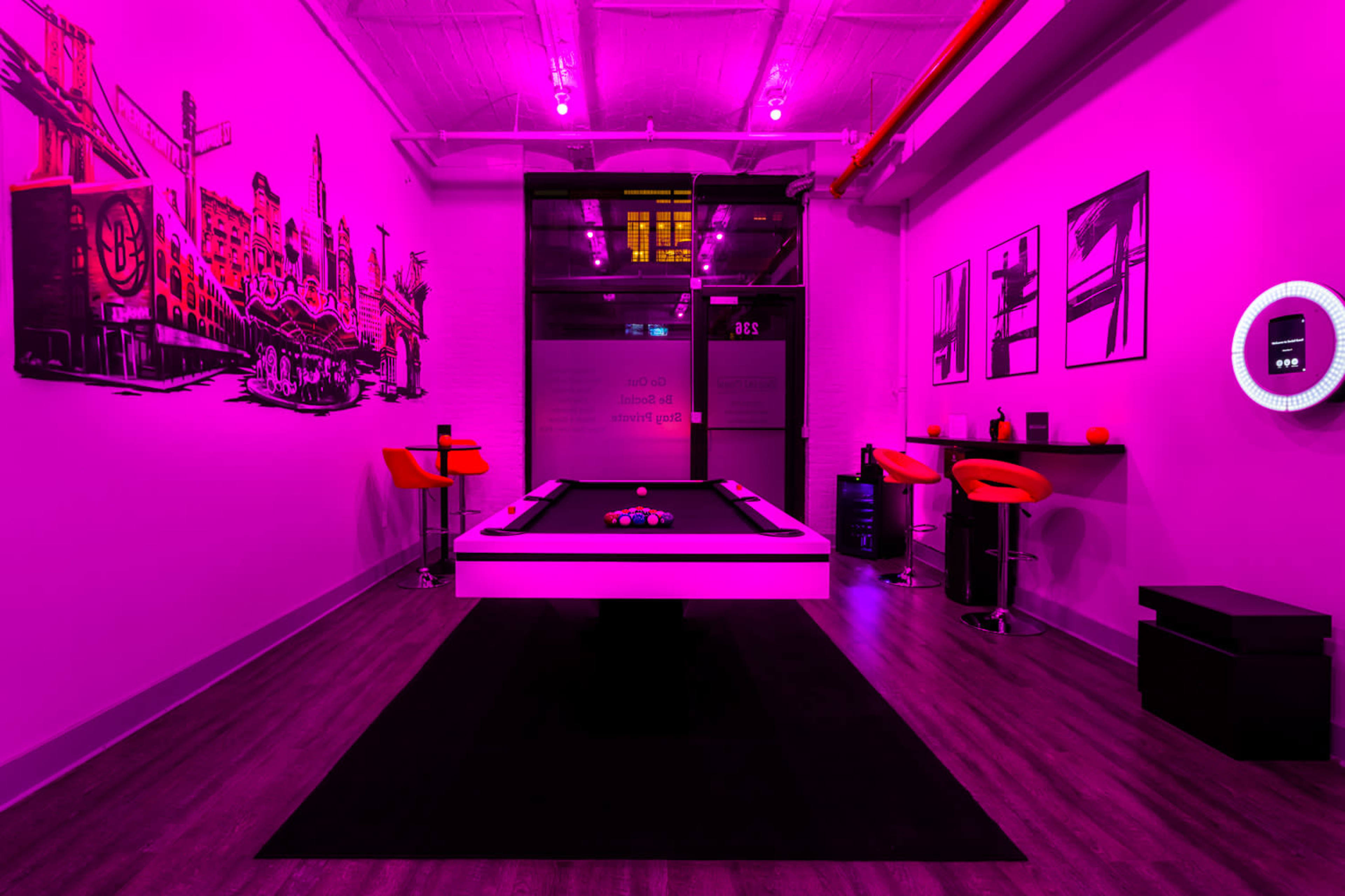 The image shows a room with bright pink walls, a modern pool table in the center, and orange bar stools against the walls.