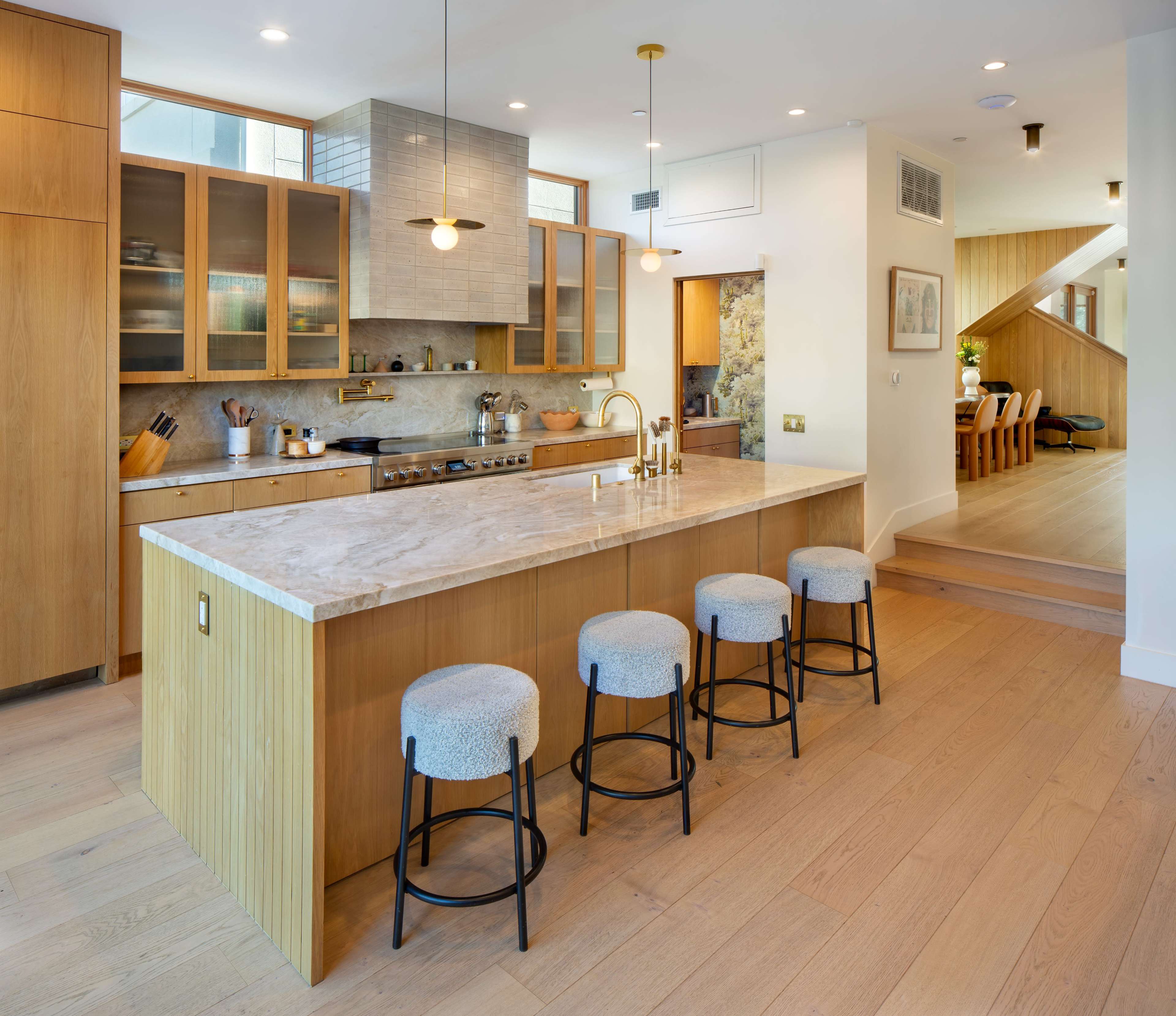 The image shows a modern kitchen with a central island, wooden cabinetry, and four stools arranged around the island.