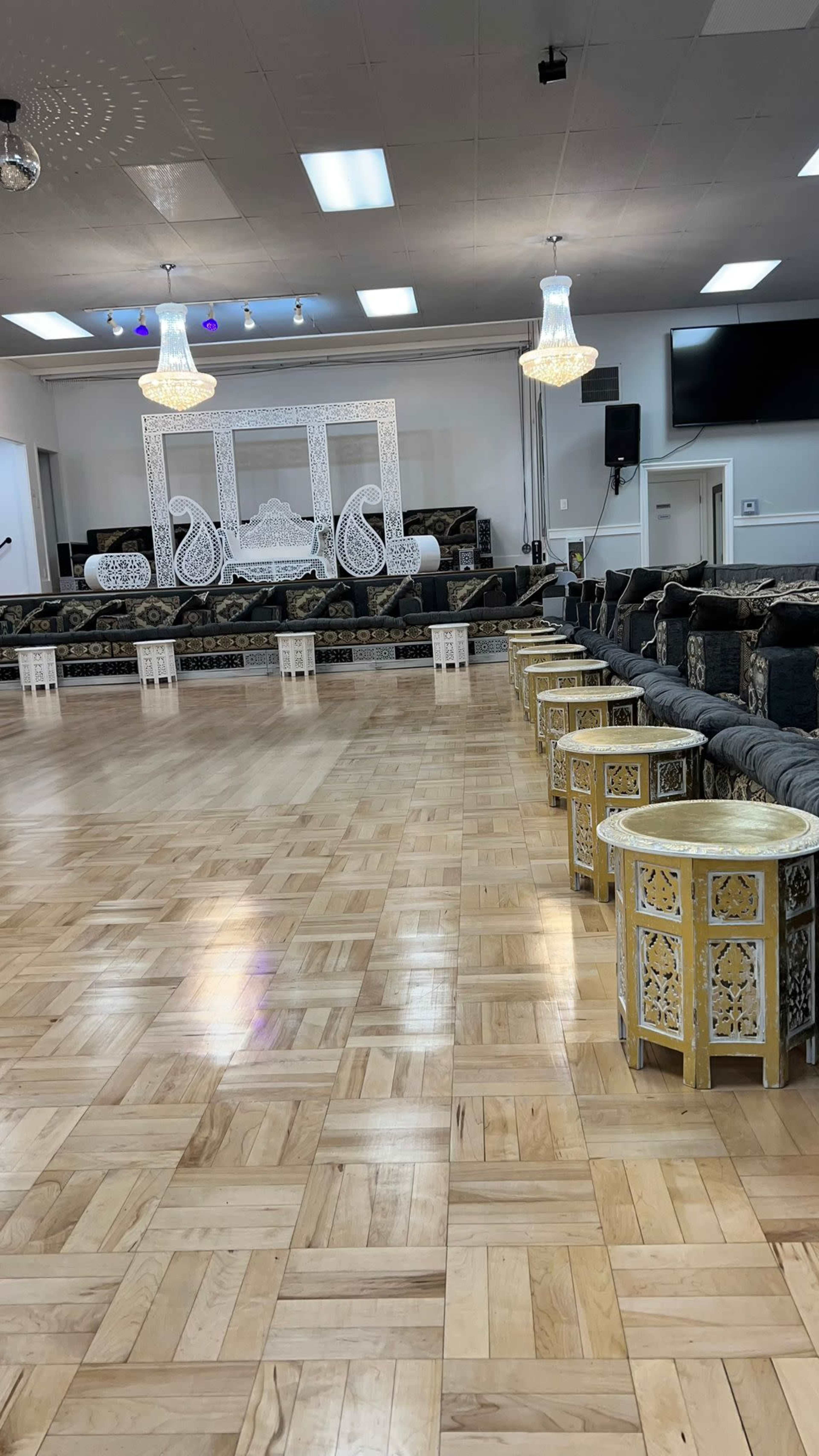 A spacious indoor venue with a polished wooden floor, intricately designed low seating areas, and decorative elements, including two large chandeliers and a white ornate backdrop.