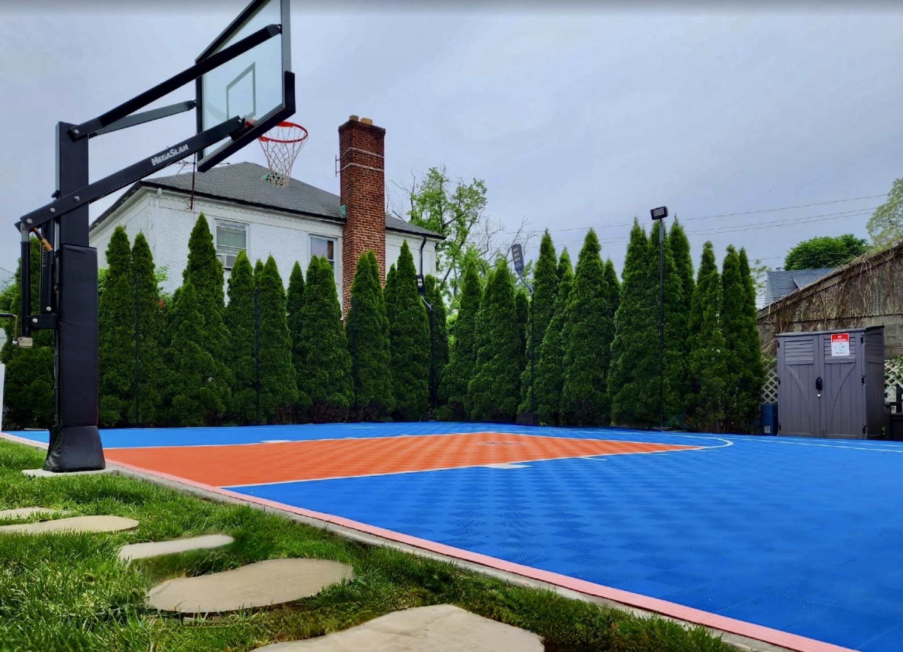 A residential basketball court with a blue and orange surface, surrounded by tall green trees and a shed in the background.