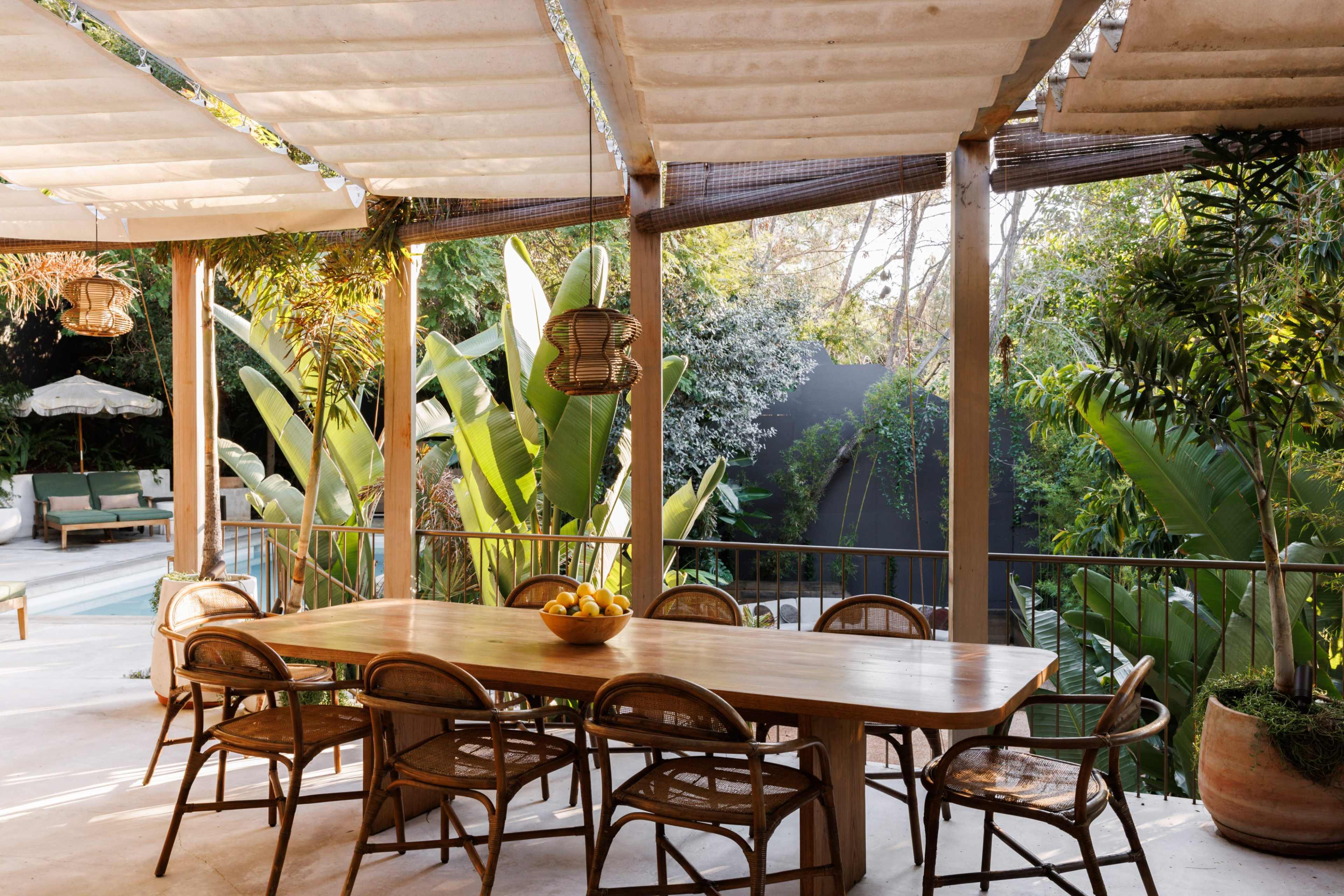 A wooden dining table surrounded by chairs is set on a patio overlooking lush greenery.