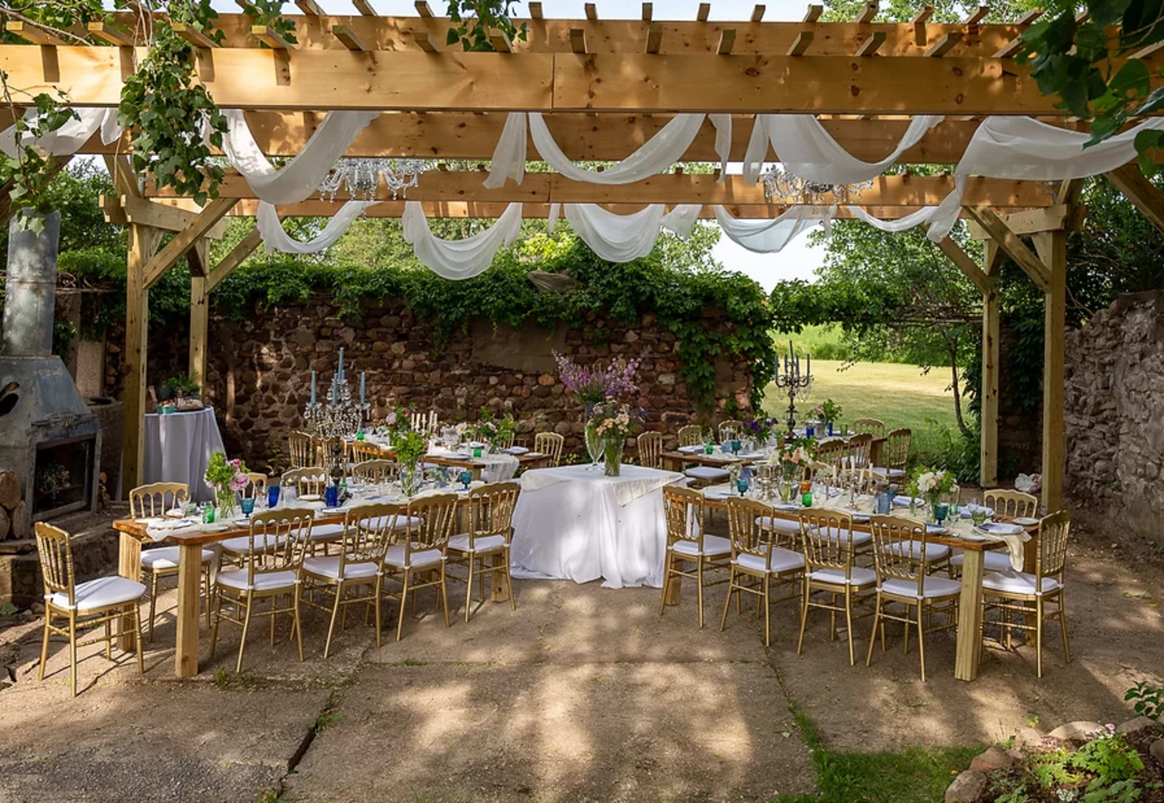 A decorated outdoor dining area features long tables with gold chairs, surrounded by greenery and draped fabric overhead.
