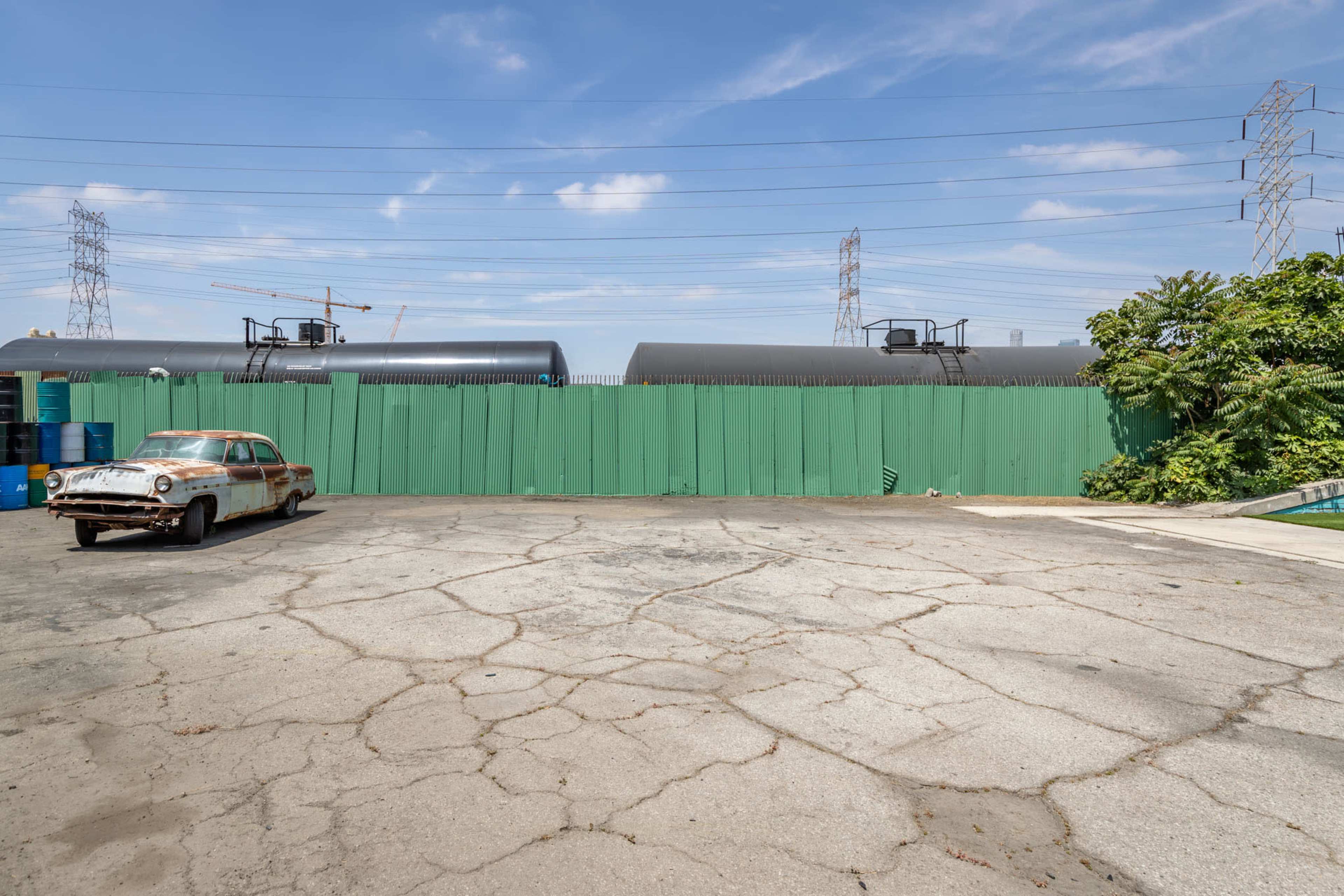 A cracked pavement area is shown with an abandoned car and a tall green fence, backed by industrial structures and power lines under a clear sky.
