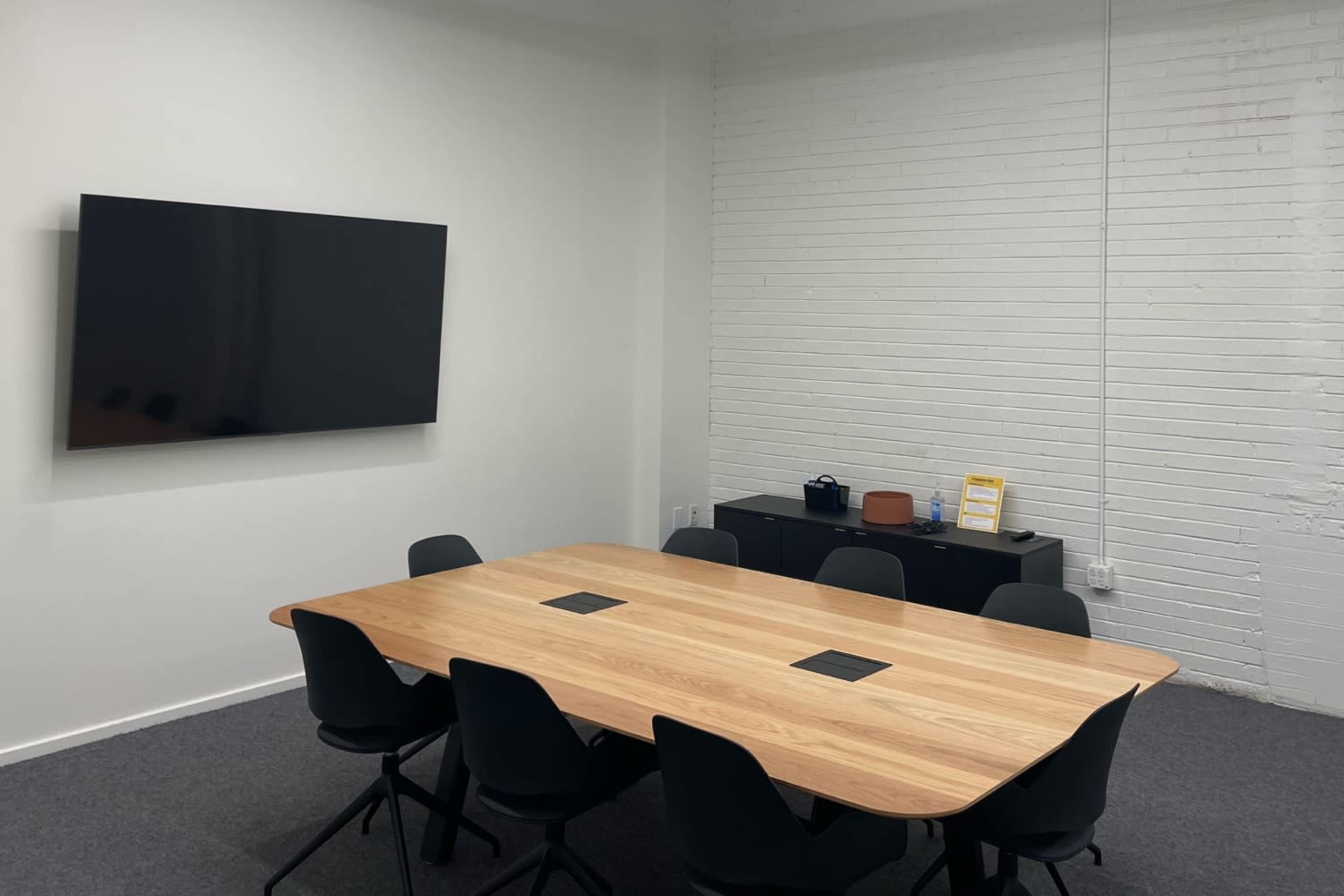 A meeting room features a long wooden table surrounded by black chairs, with a wall-mounted television and a low black cabinet.