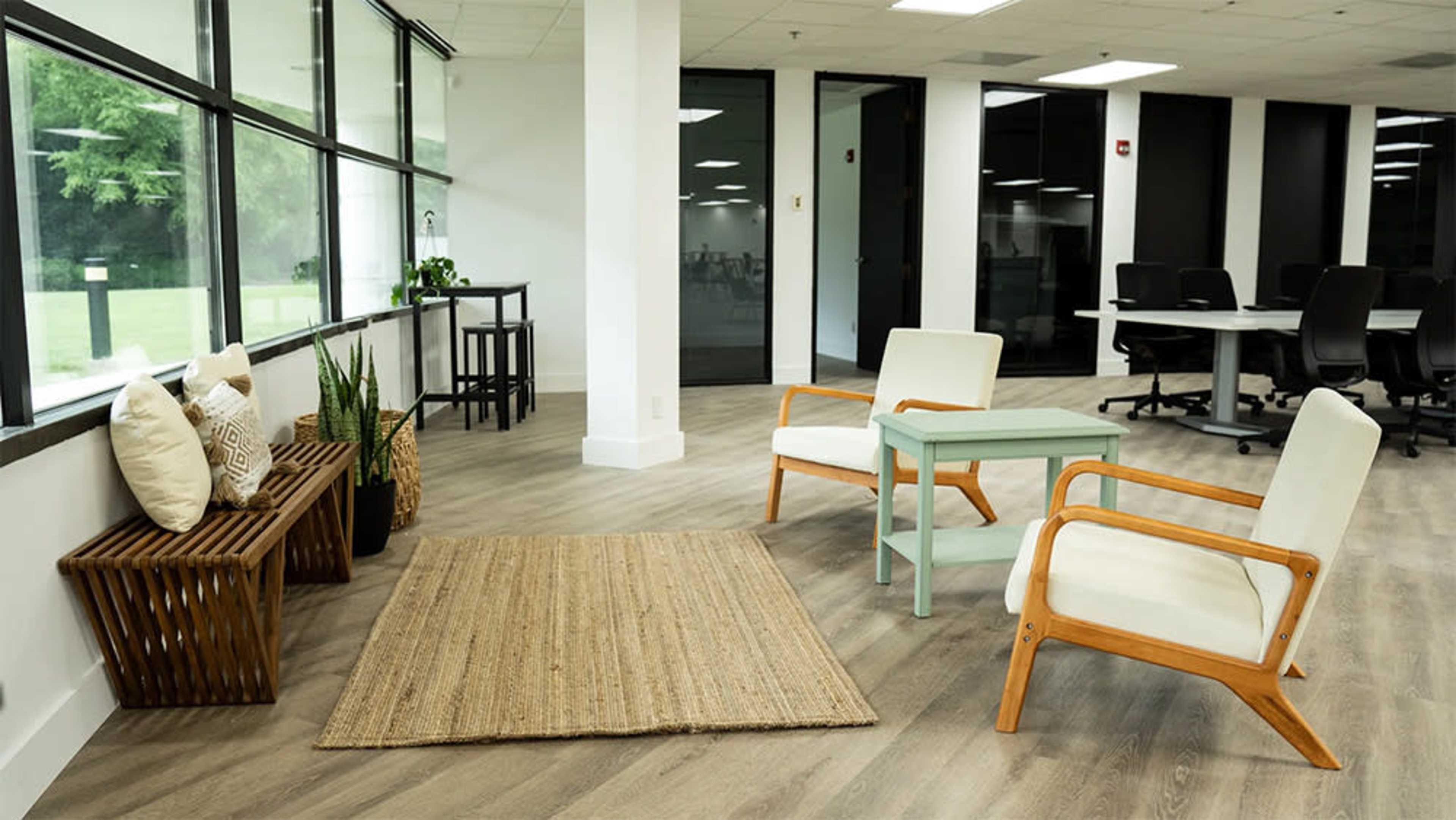 A modern office lounge area with two light-colored chairs, a small green table, a woven rug, and a wooden bench adorned with pillows.