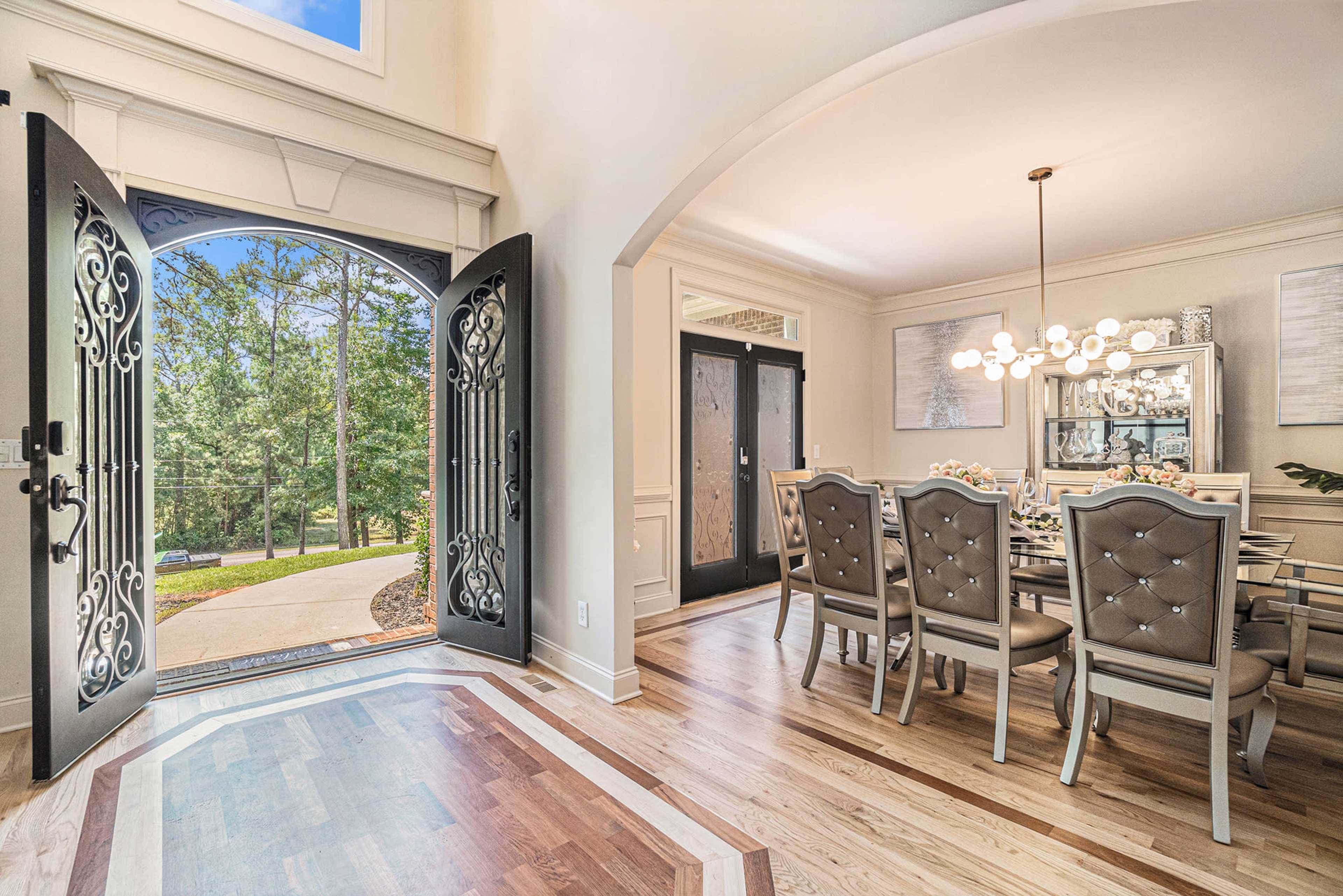 The image shows a large, open entryway leading to a dining room with a view of lush greenery outside through double doors.